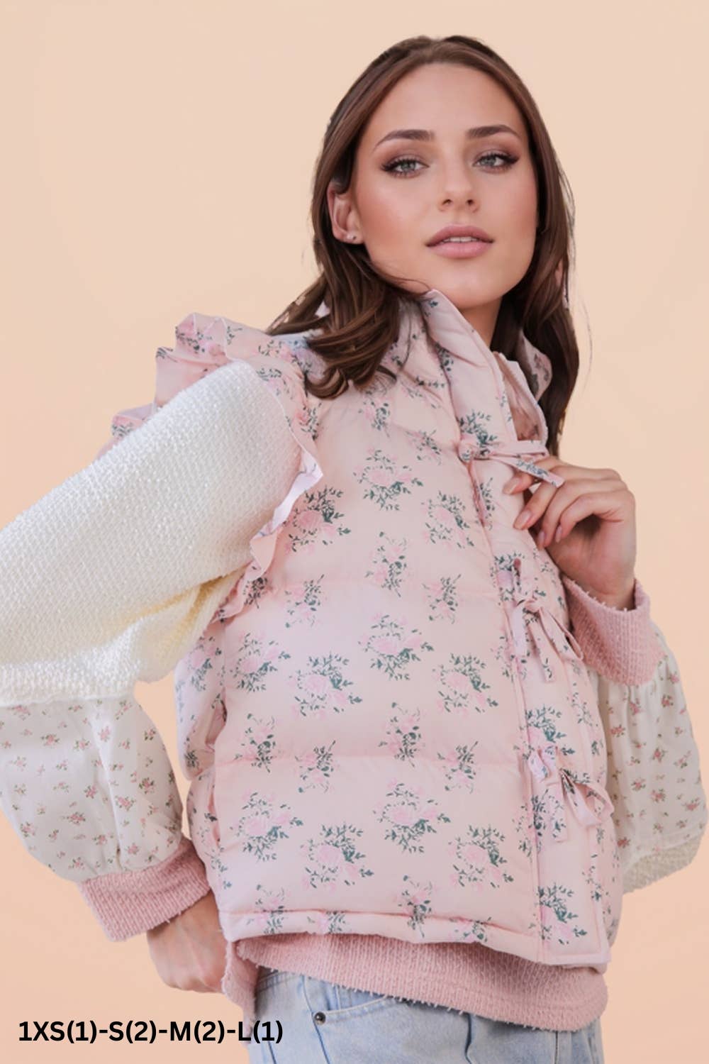 Woman wearing a pink floral puffer vest  with a white cardigan on a peach background