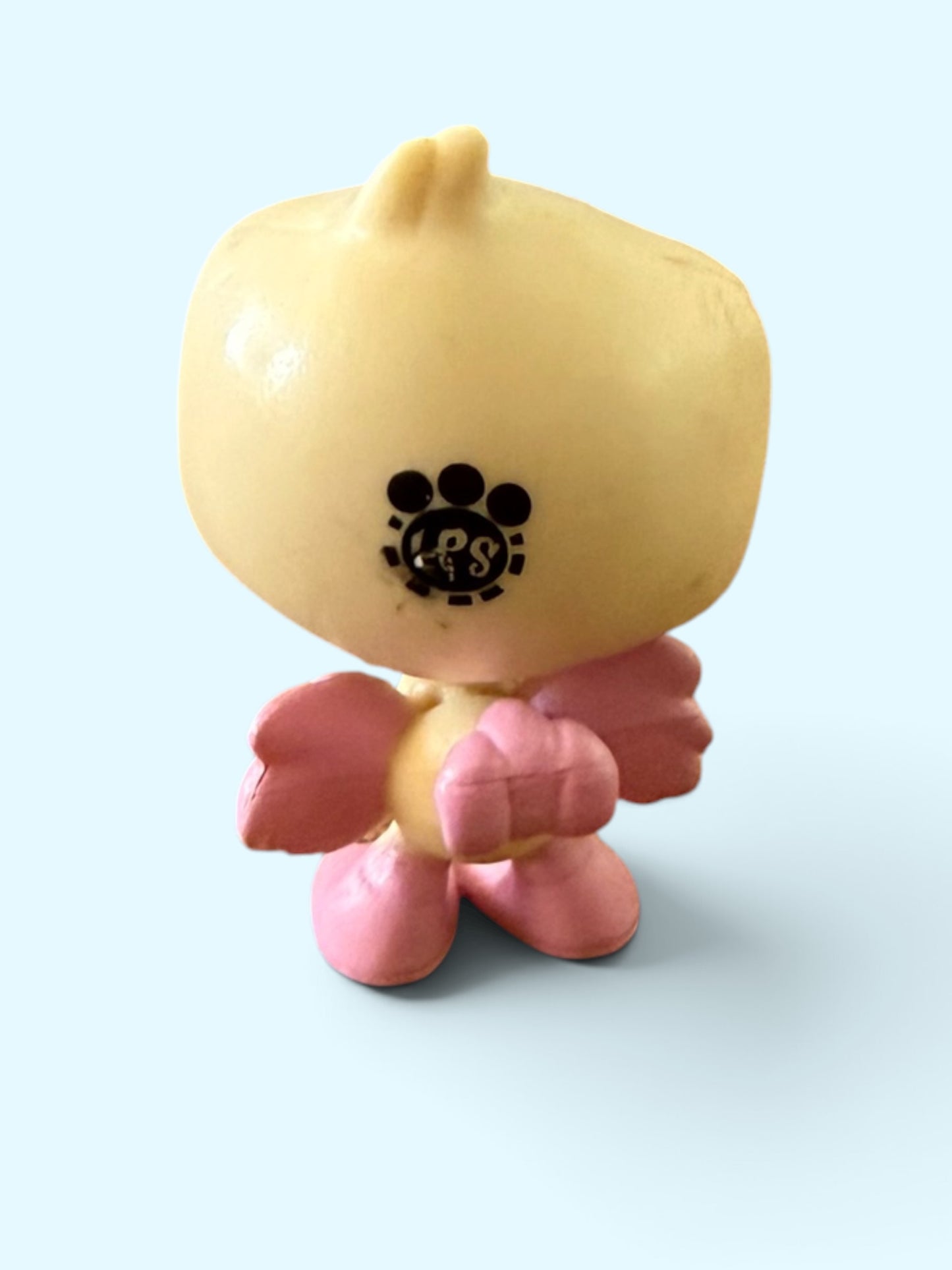 A small stylized toy figure with a rounded pale yellow head and body, pink wings and feet, standing against a light blue background. A black paw-print style logo with the letters 'PS' is printed on the front of the head.