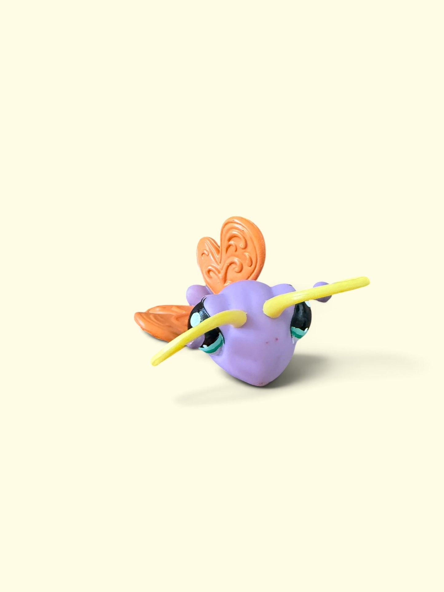 A small figurine of a purple creature with orange wings and yellow antennae, set against a solid pale cream background.