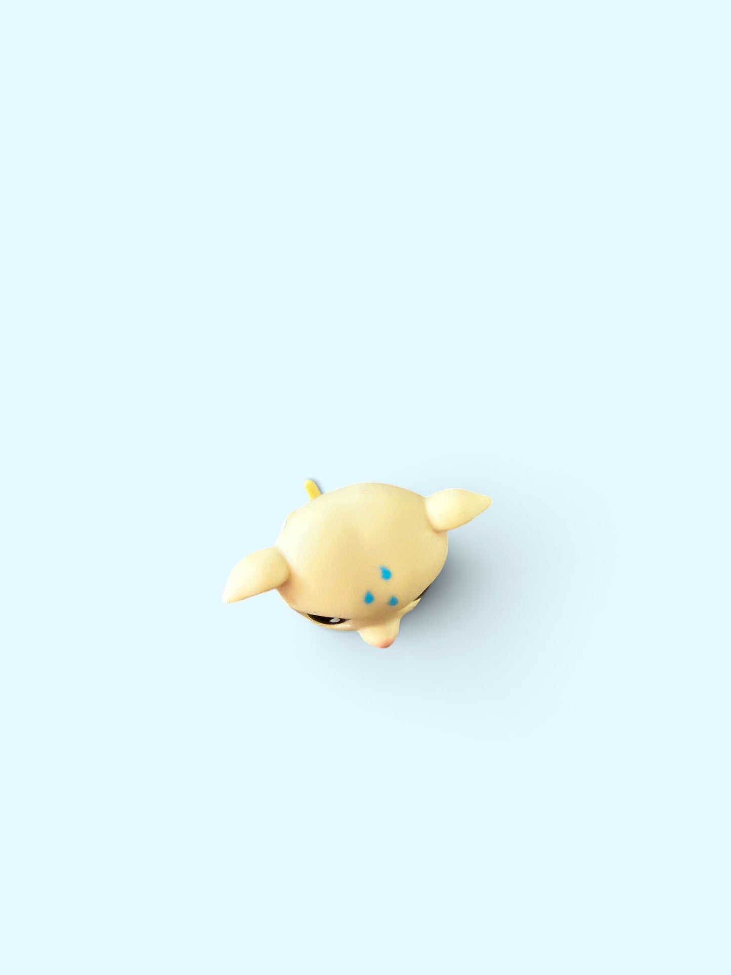 A small pale yellow cartoon figurine with blue speckles and a black eye, shown from above on a light blue background.