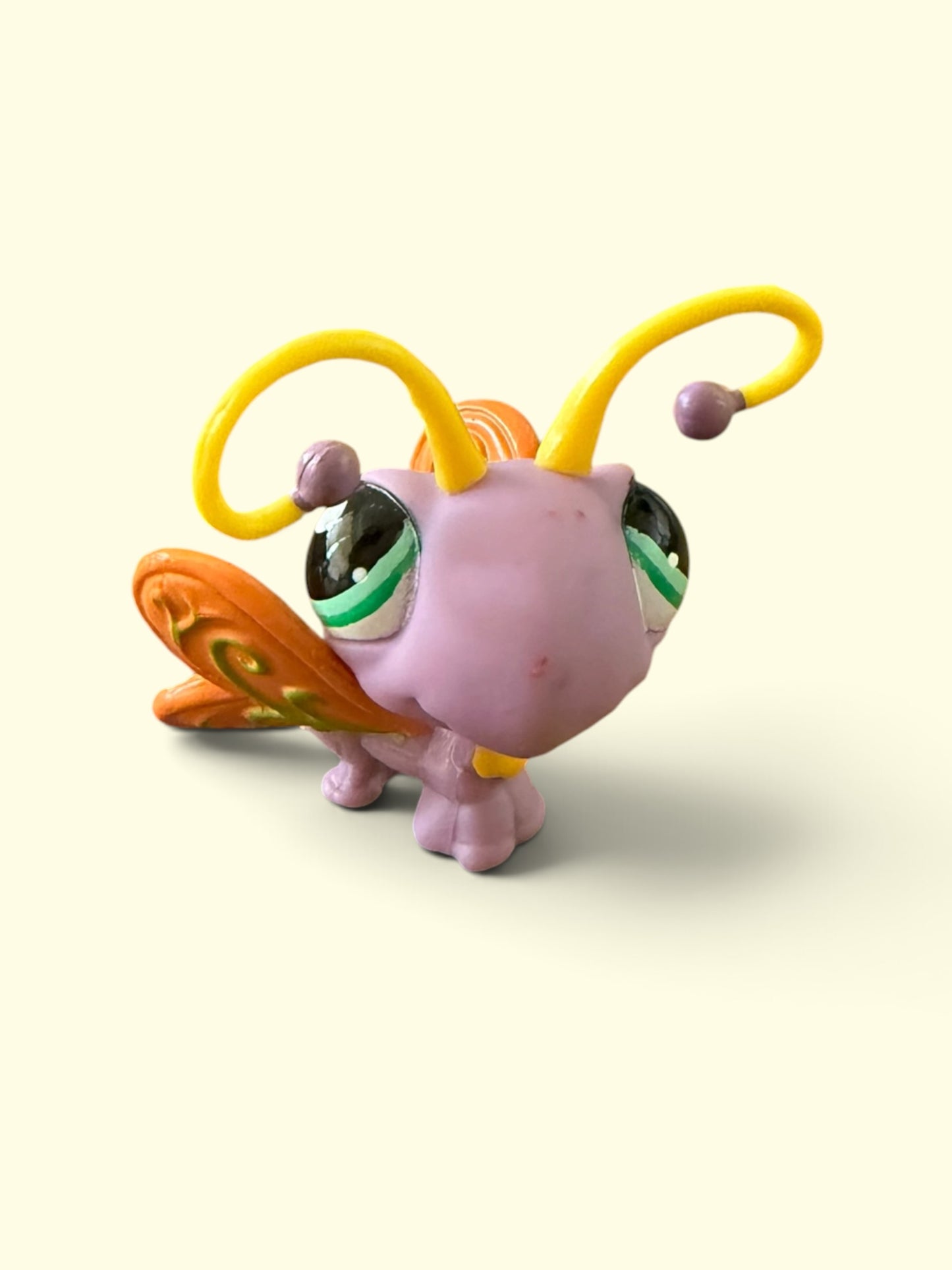 A small plastic toy figure shaped like a butterfly or beetle, with a pink body, large green and black eyes, yellow curved antennae, and orange patterned wings, photographed on a plain light cream background.