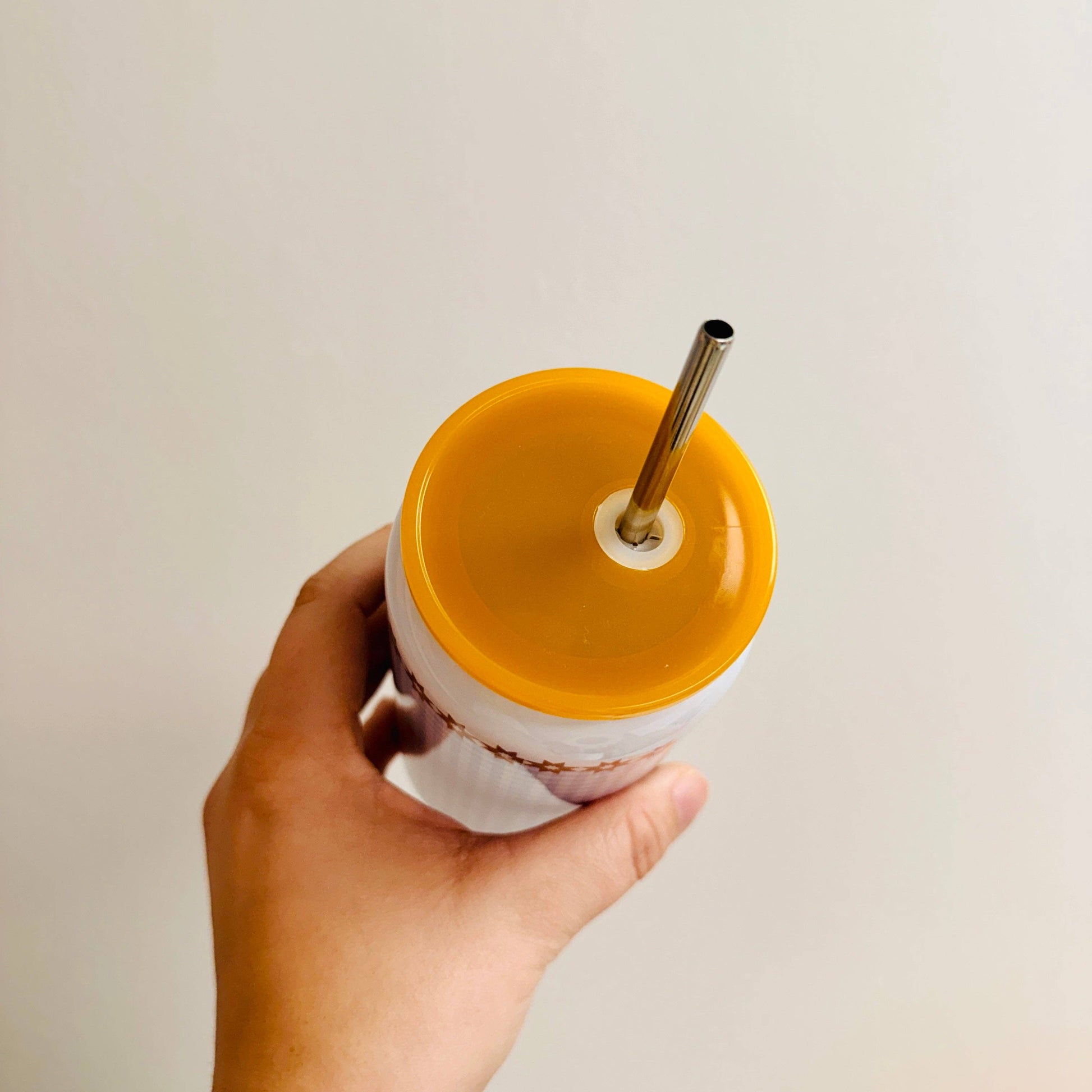Hand holding a white cup with a yellow lid and metal straw against a plain background