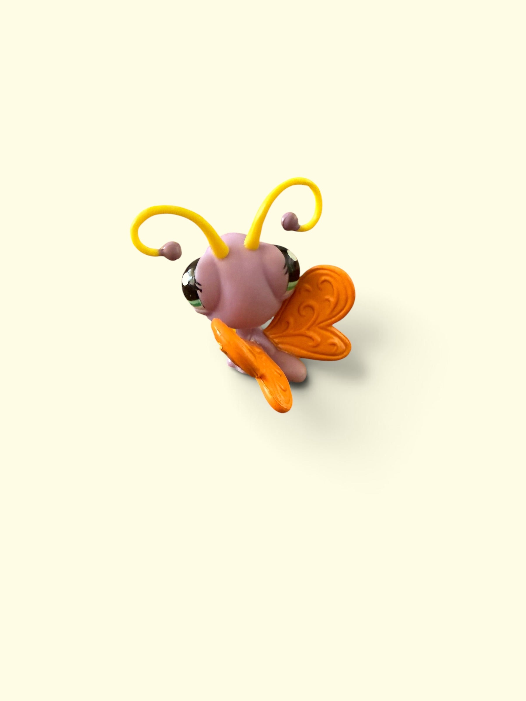 A small stylized bee figurine with a pink body, yellow antennae, and orange wings, shown on a plain warm cream background.