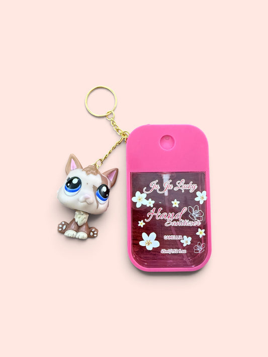 A pink hand sanitizer bottle with a small cat figurine keychain attached, set against a solid pale pink background. The bottle features a clear window showing a dark pink liquid with white text that reads 'Just For Lady Heart-shaped Sanitizer' and 'GIRLS ONLY'.