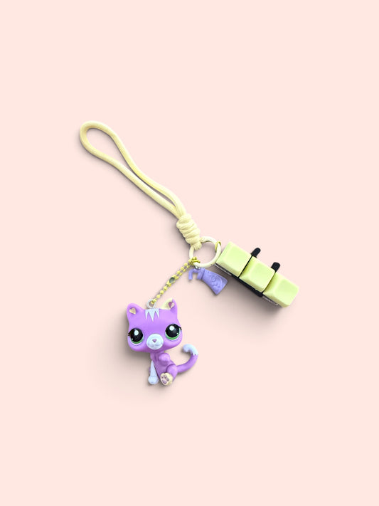 A pastel-colored keychain or bag charm featuring a purple cat figure, a light green rectangular charm, and a pale yellow loop strap, set against a solid pale pink background.