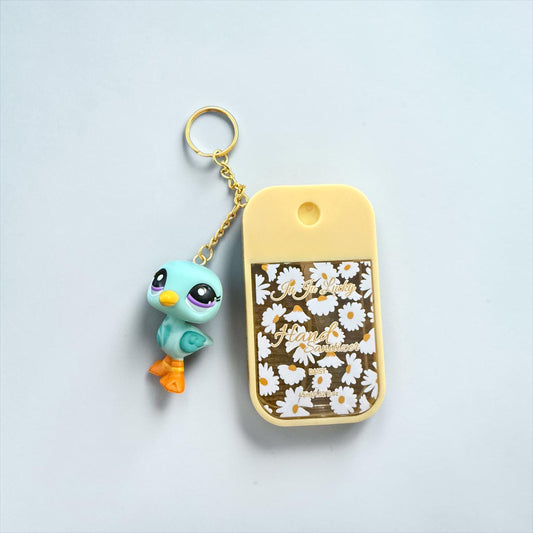 A small keychain accessory set on a light gray background, featuring a turquoise and orange bird charm attached by a gold chain to a yellow rectangular card case with a white daisy floral print and the word 'Peach' in gold script.