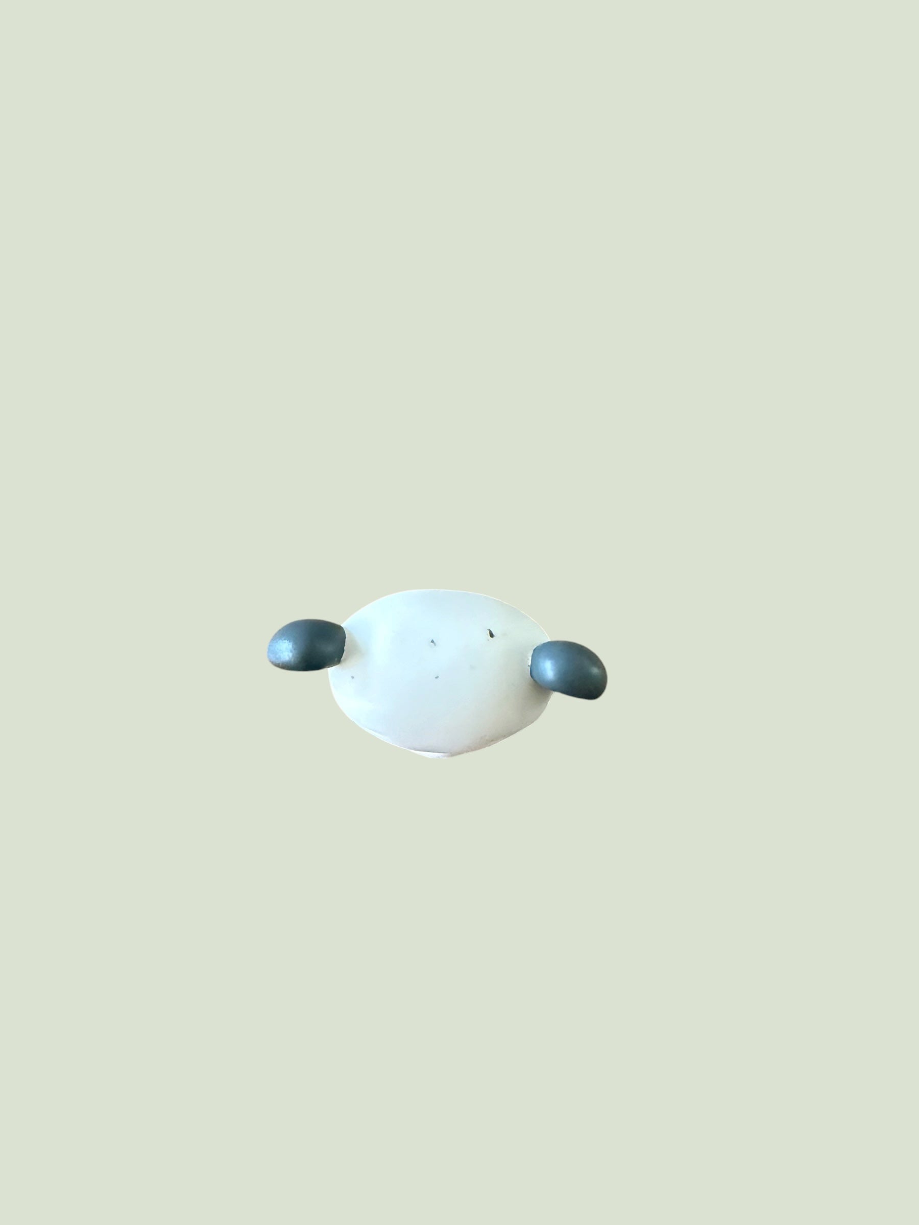 A minimalist abstract wall art piece featuring a smooth, rounded white central oval with two small, dark slate-gray teardrop shapes on either side, set against a solid pale sage green background.