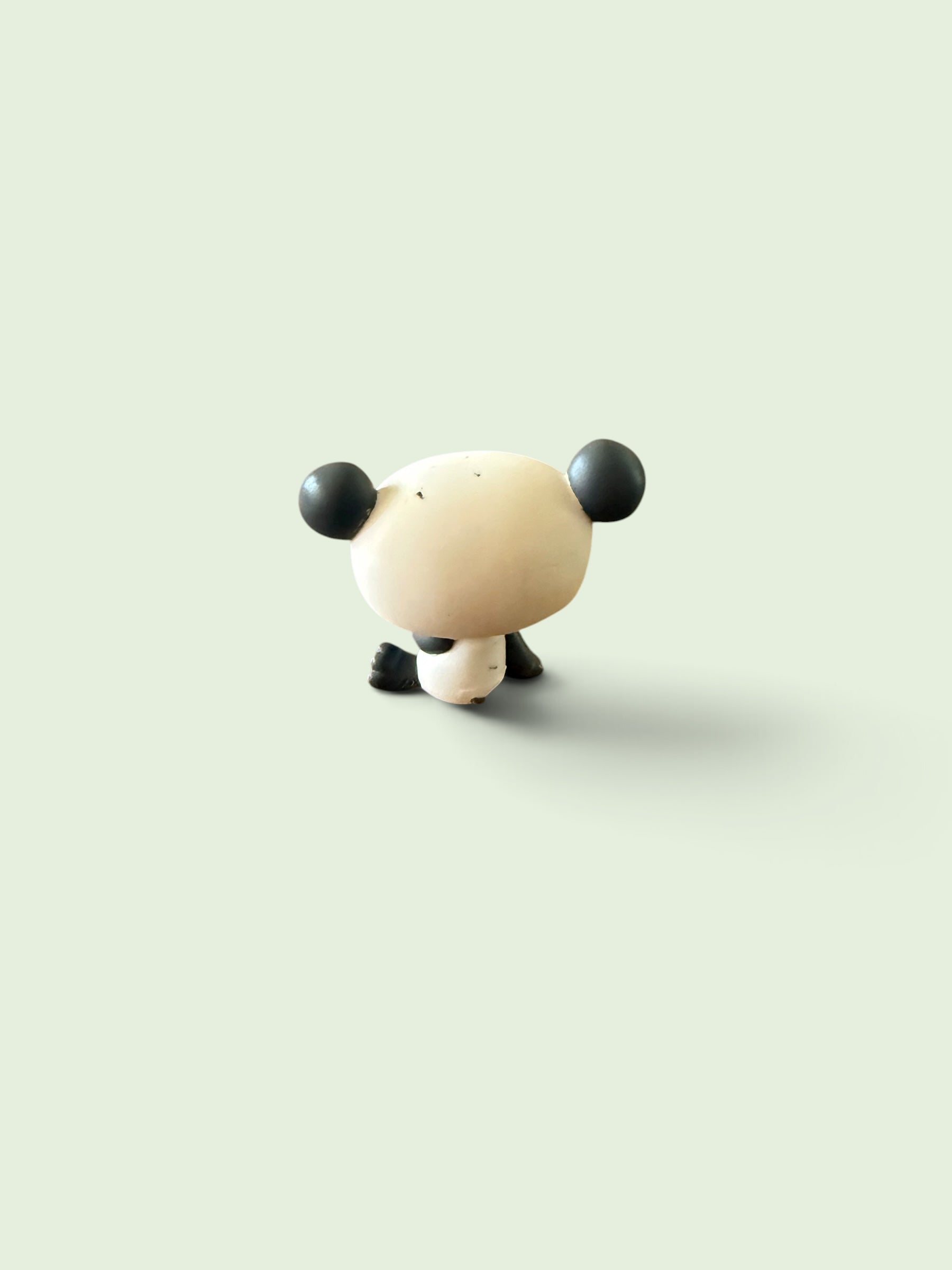 A small stylized panda figurine viewed from the back, with a cream-colored body and black ears, feet, and lower body, centered on a pale green background.