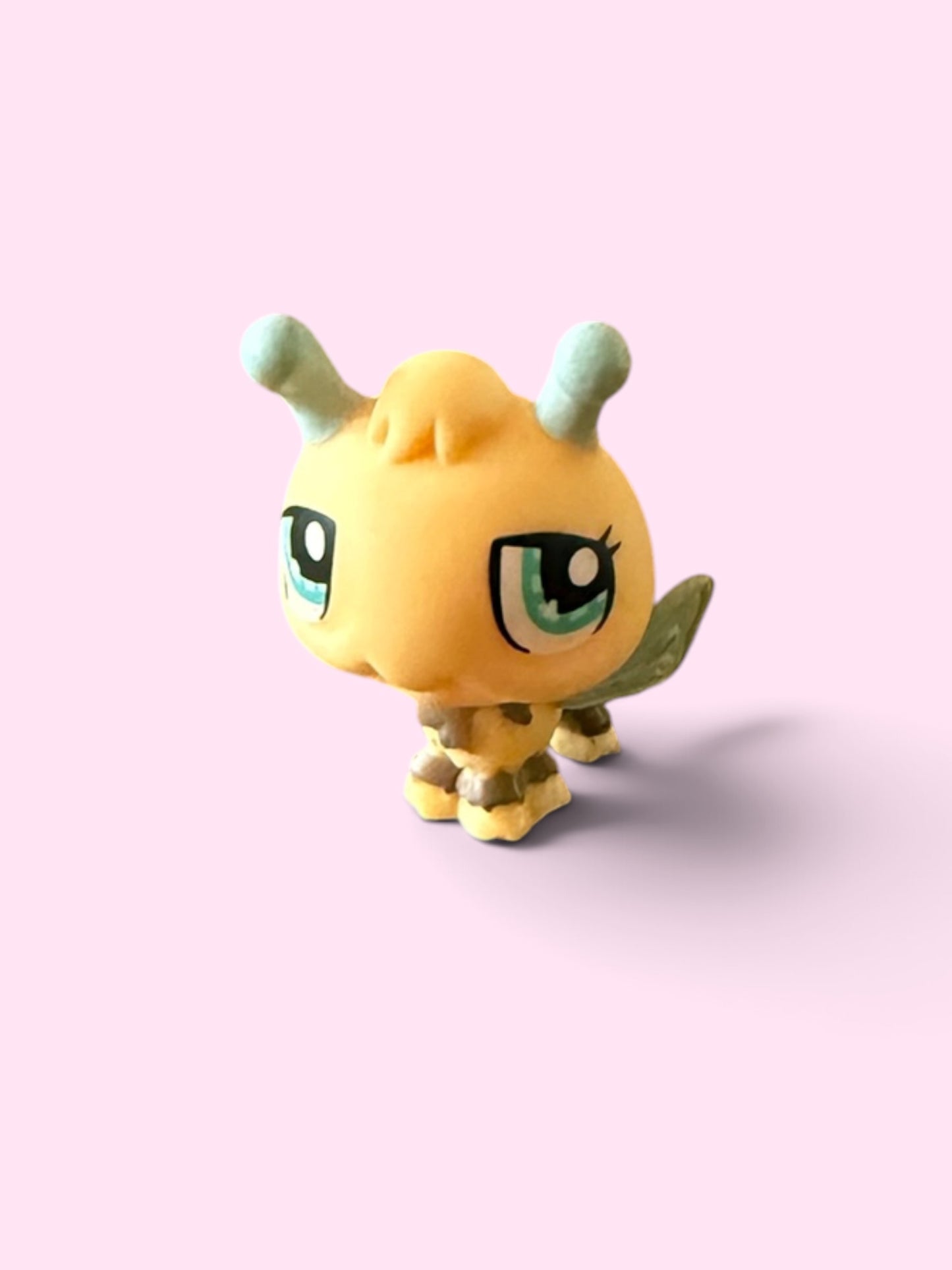 A small yellow cartoon creature figurine with large green eyes, mint green horns, and gold stripes, standing on a light pink background.