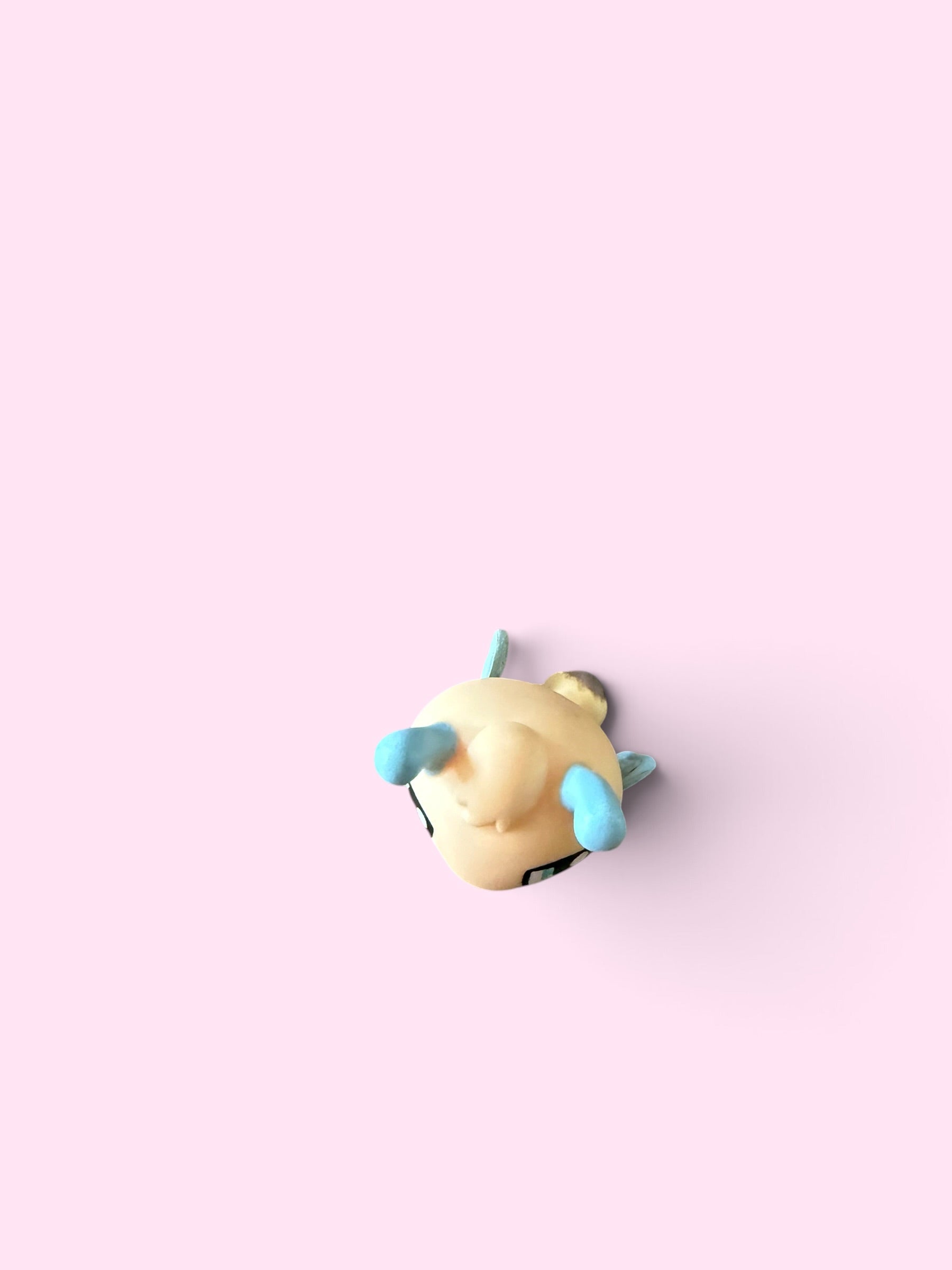 A small cream-colored plush toy with light blue ears and a gold bell, photographed from above on a solid pastel pink background.
