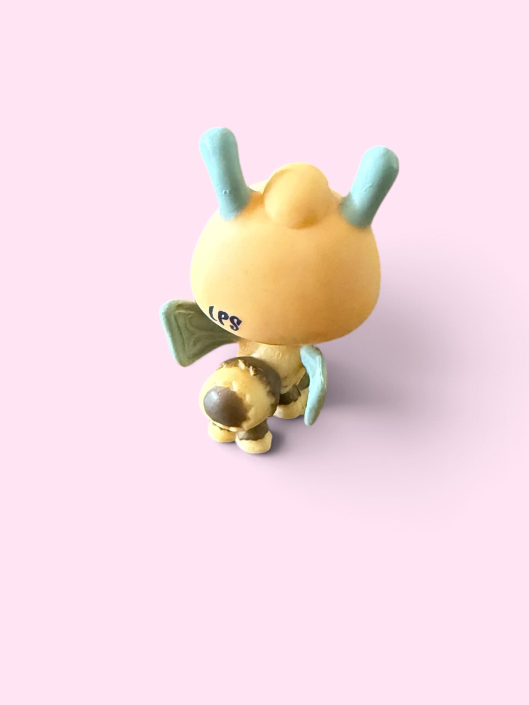A stylized yellow bunny figurine with light blue ears and wings, shown from the back on a solid light pink background.