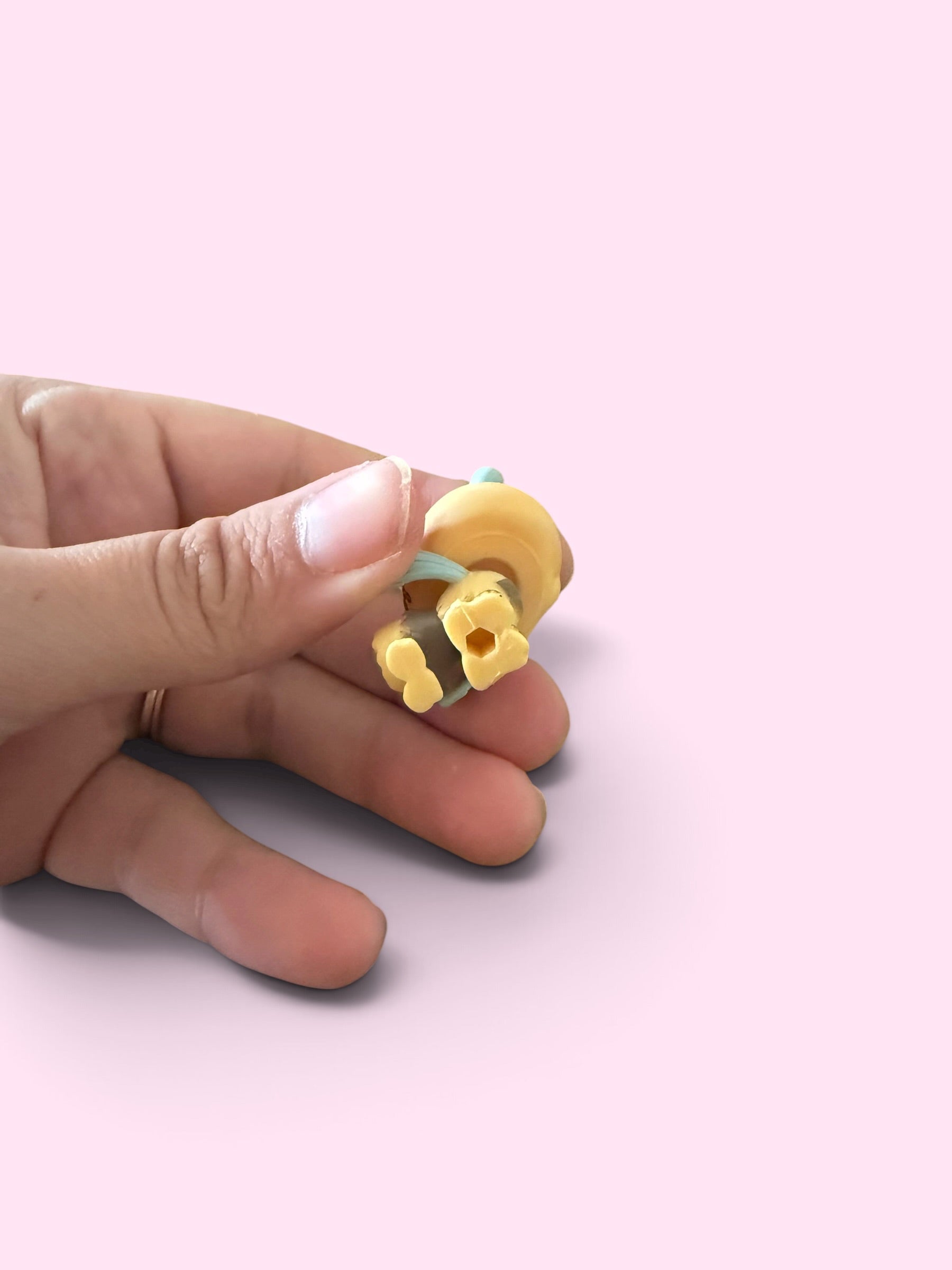 A hand holding a small yellow and mint green ring-shaped accessory against a solid light pink background.
