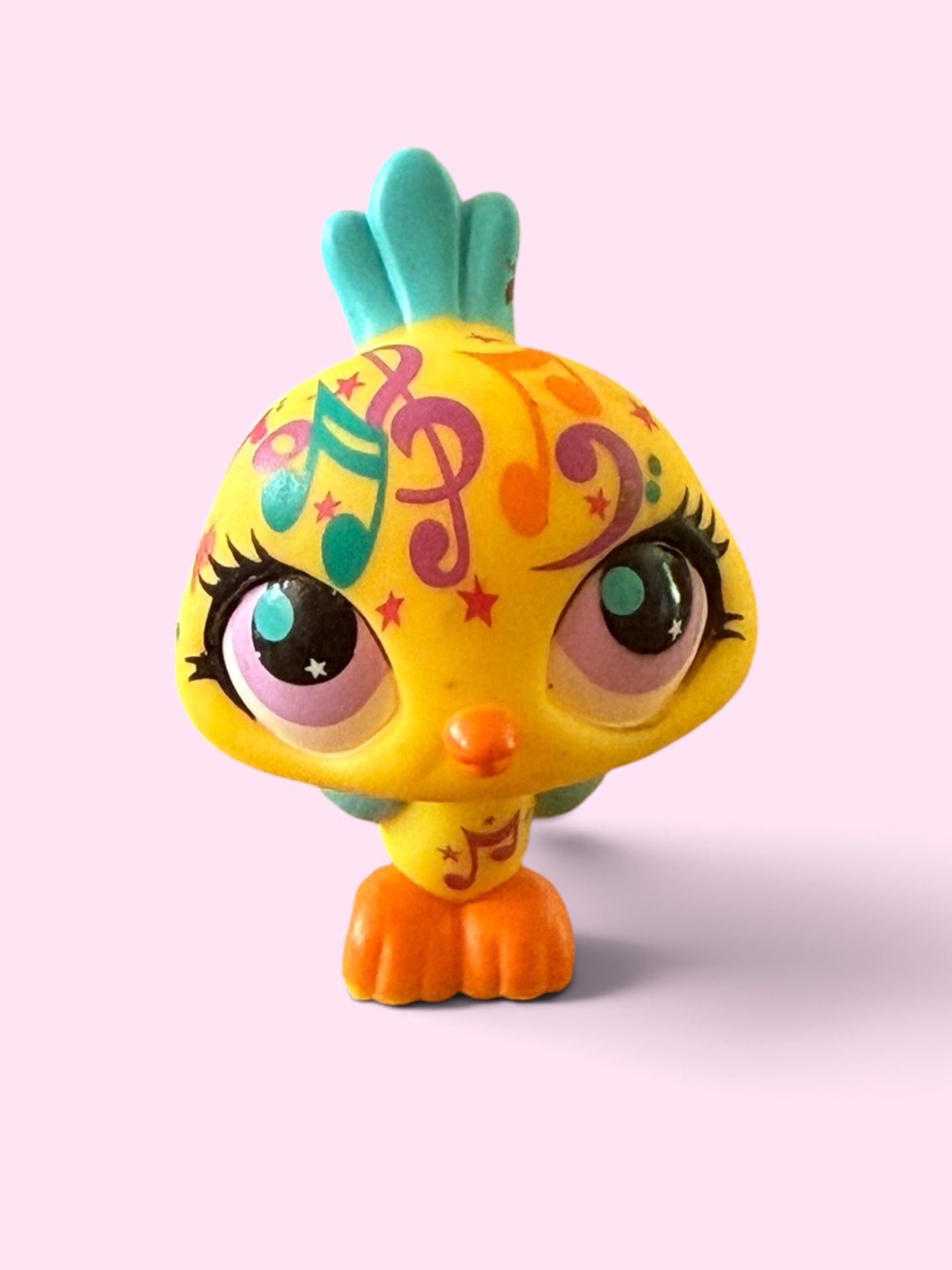 A small stylized bird figurine with a bright yellow body, orange feet and beak, teal tail feathers, and multicolored decorative swirls and stars on its head, centered on a pale pink background.