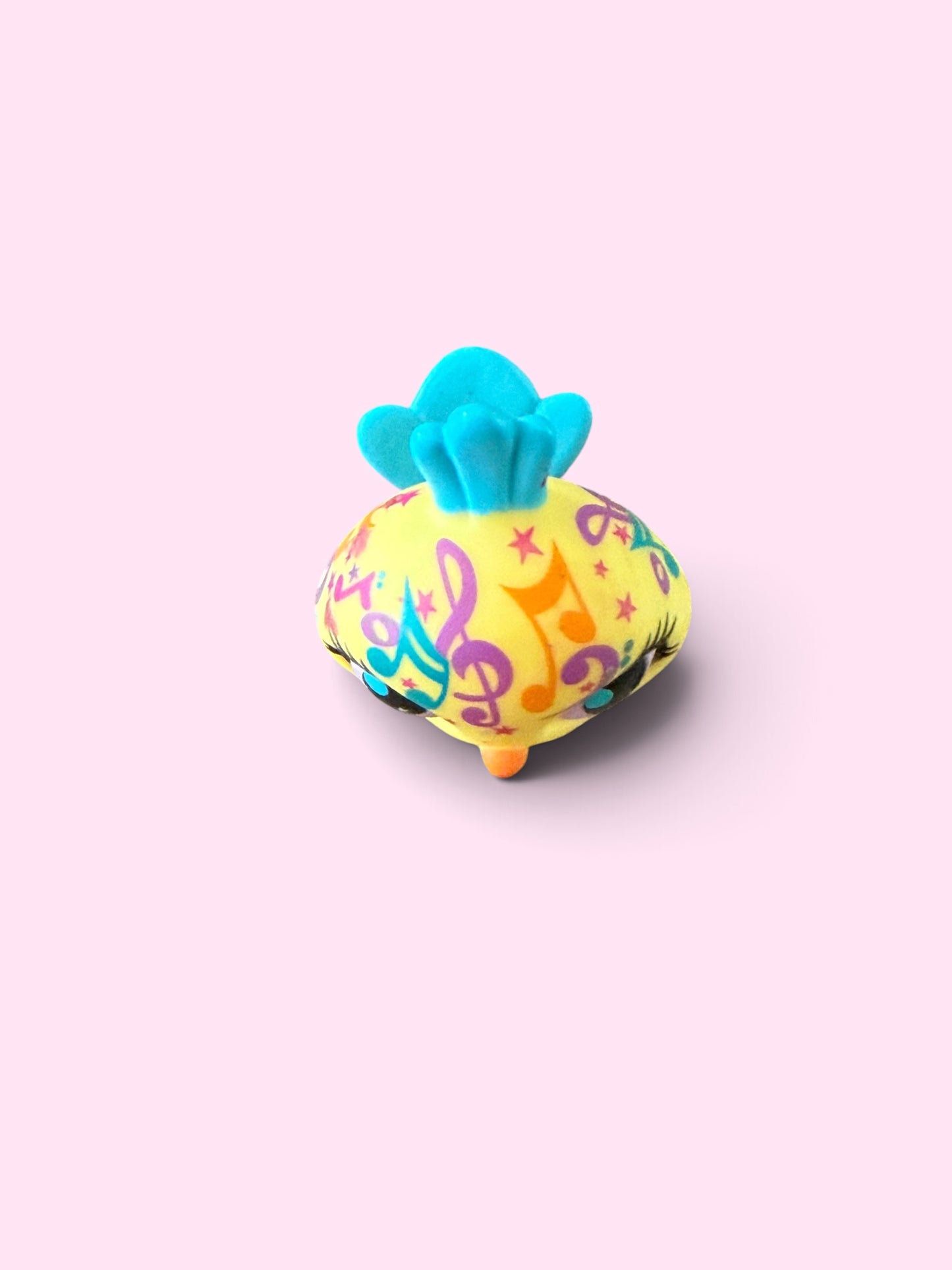 A small decorative cushion or pillow with a bright yellow body covered in colorful musical note and star patterns, topped with a turquoise blue ruffled fabric, shown on a solid pale pink background.