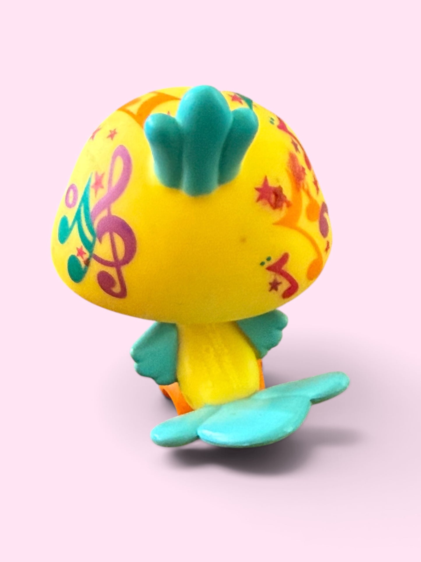 A small stylized toy fish or mermaid figure in bright yellow with turquoise fins and tail, decorated with colorful musical notes and stars, shown on a soft pink background.