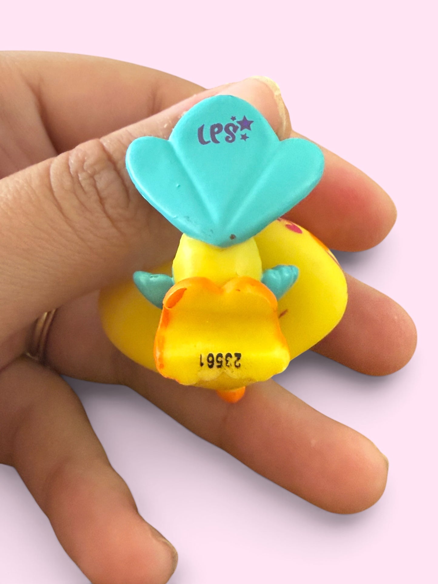 A small yellow and turquoise toy figure being held between fingers against a pale pink background. The toy has a turquoise top section with the letters 'LPS' and a yellow body with orange details.