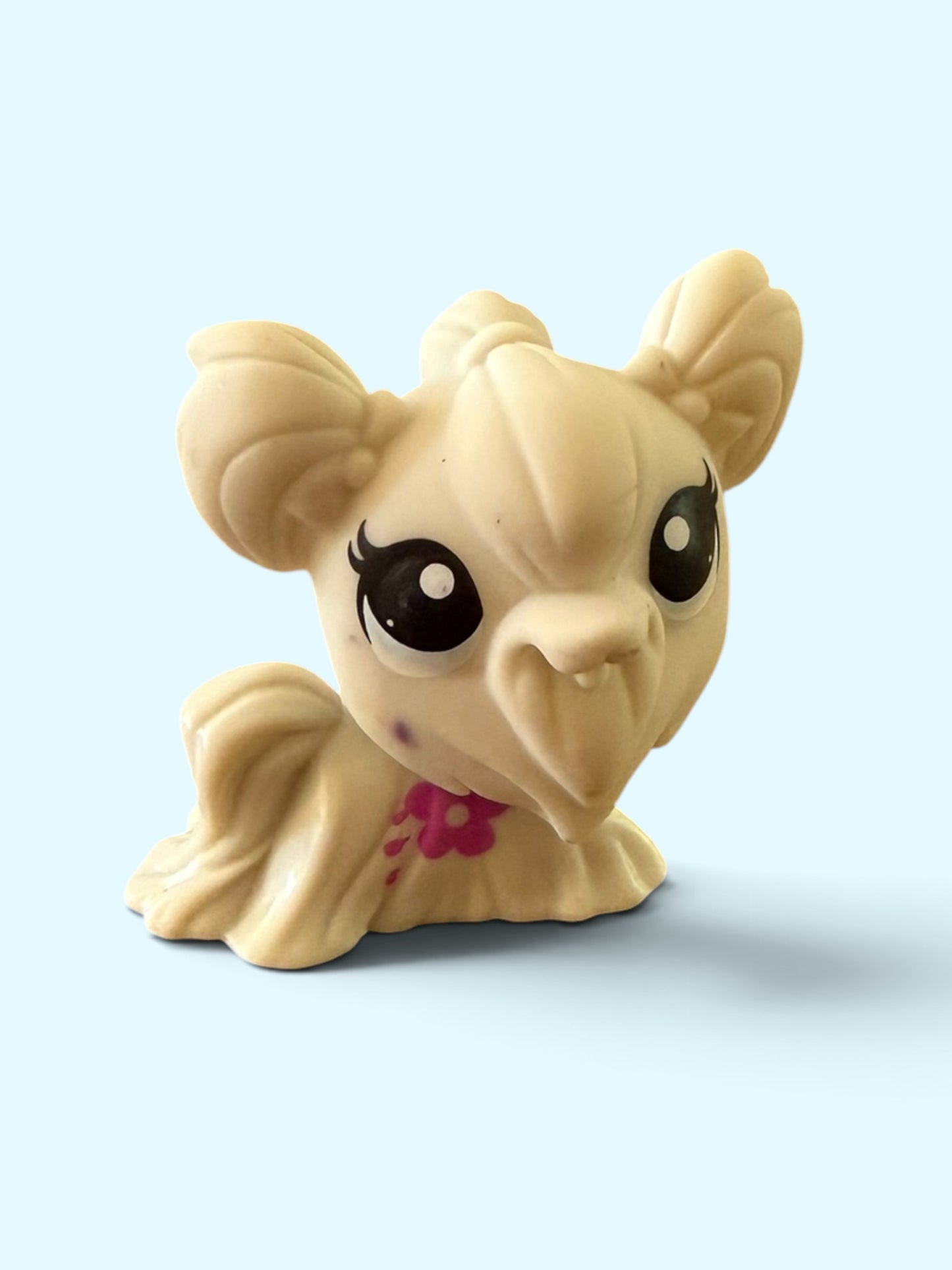 A small beige toy pony figurine with large black eyes, a pink flower detail on its neck, and a light blue background.