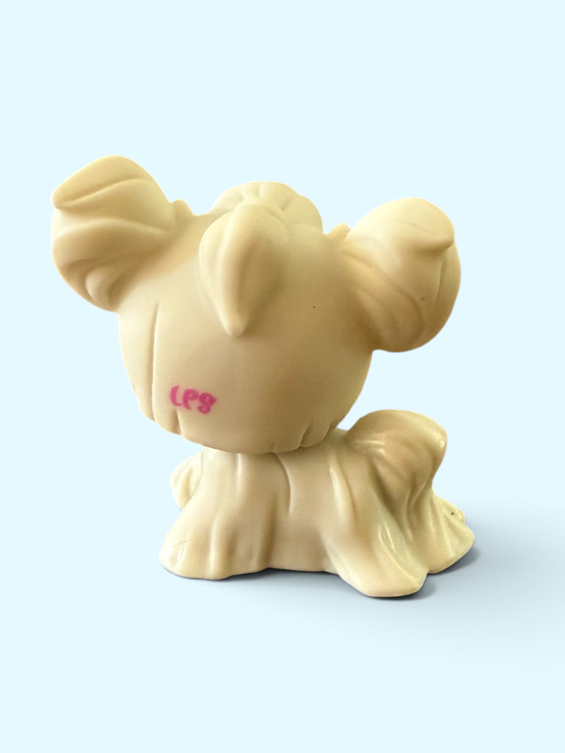 A small beige plastic toy figure with a stylized animal-like shape, shown from the back against a light blue background. The figure has pink handwritten markings near the top left.
