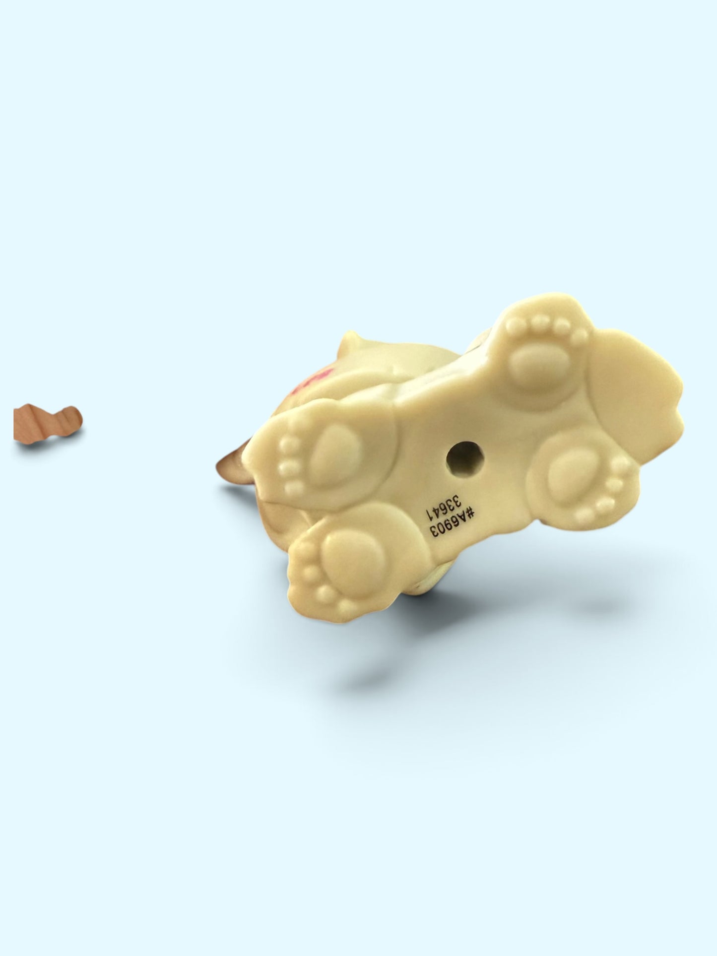 A small beige dog-shaped figurine or charm shown from the underside on a pale blue background, with a tiny brown detached piece to the left and black printed text near the feet.