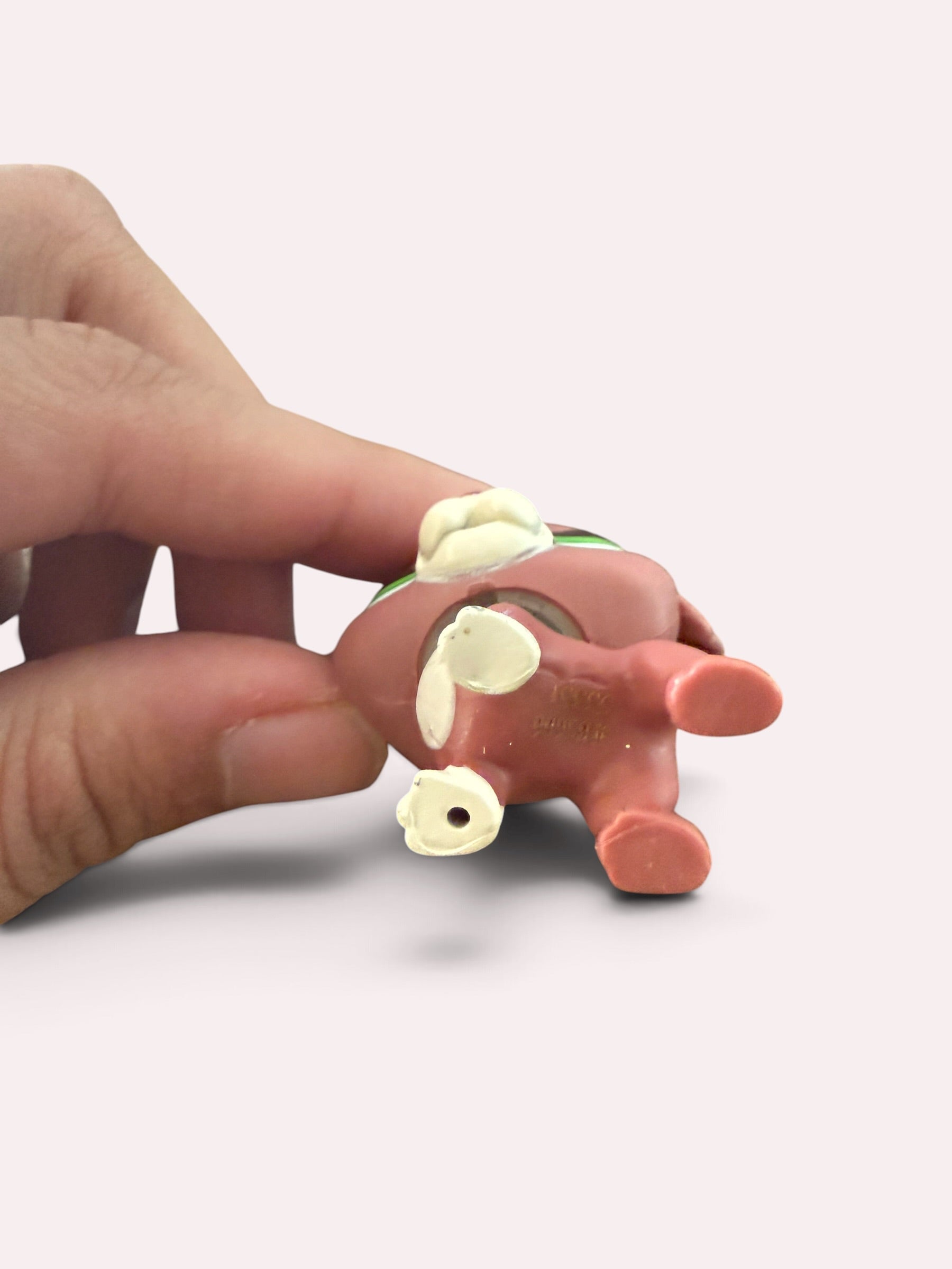 A small pinkish-brown animal figurine with cream-colored ears and tail is being held by a human hand against a plain light background.