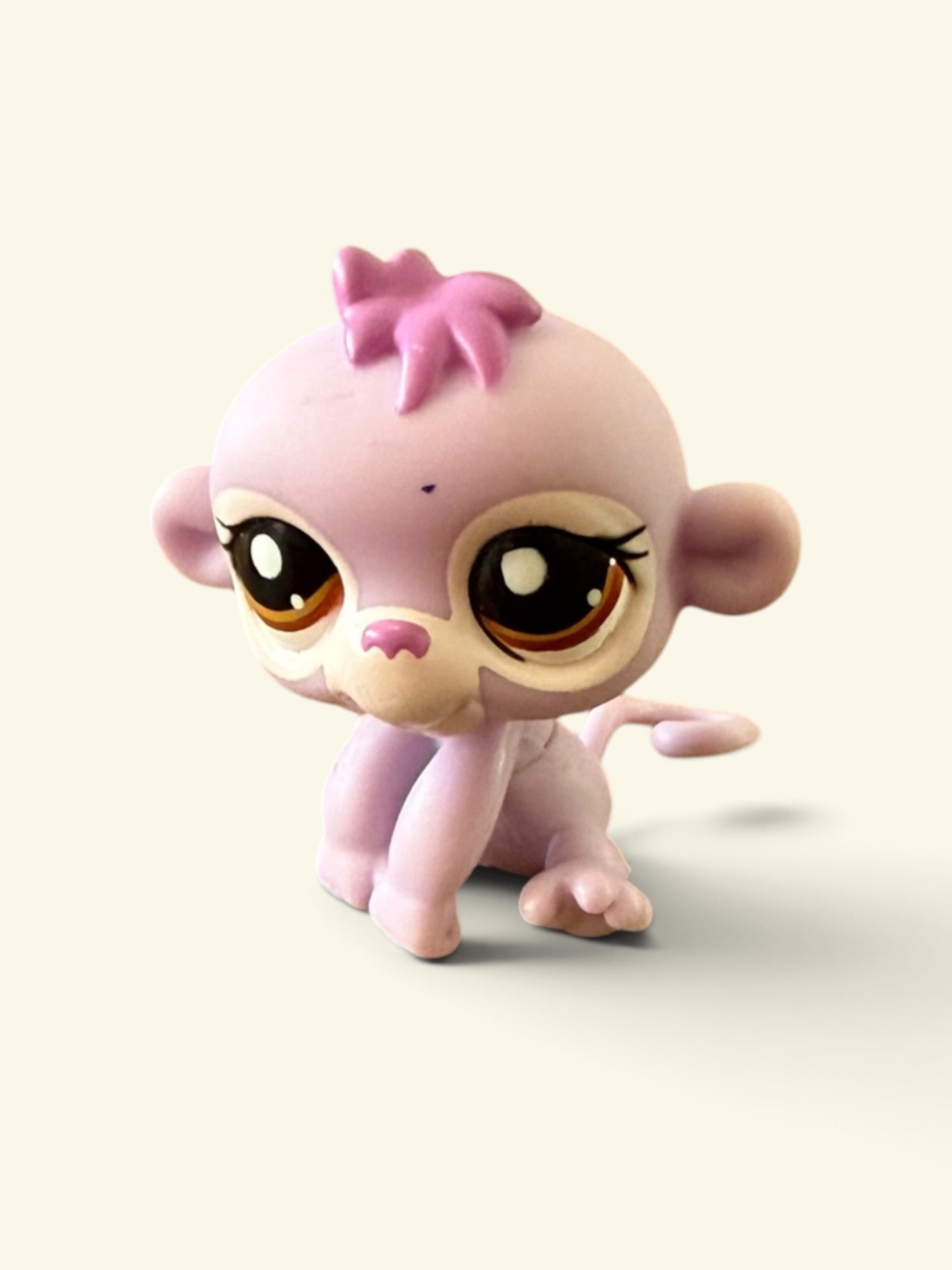 A small pink monkey toy figurine with large cartoon eyes, a pink bow on its head, and a beige muzzle, shown against a plain light cream background.
