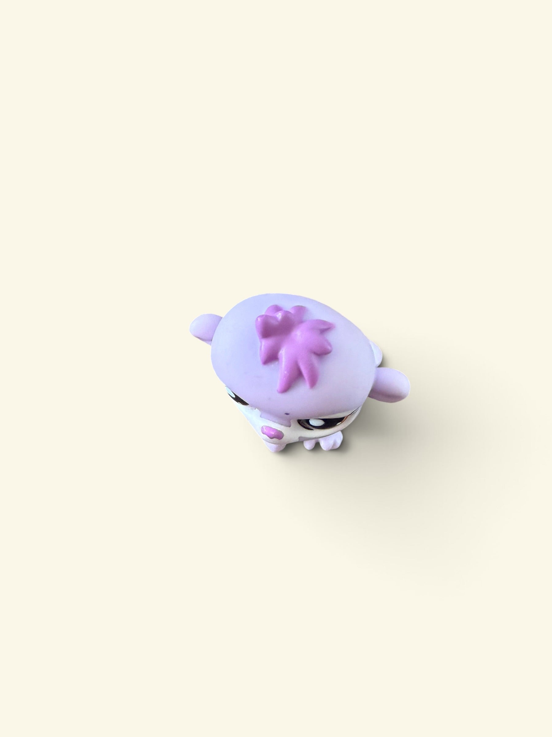 A small pastel purple and pink animal figurine, resembling a lamb or sheep with a flower on its head, centered on a solid pale cream background.