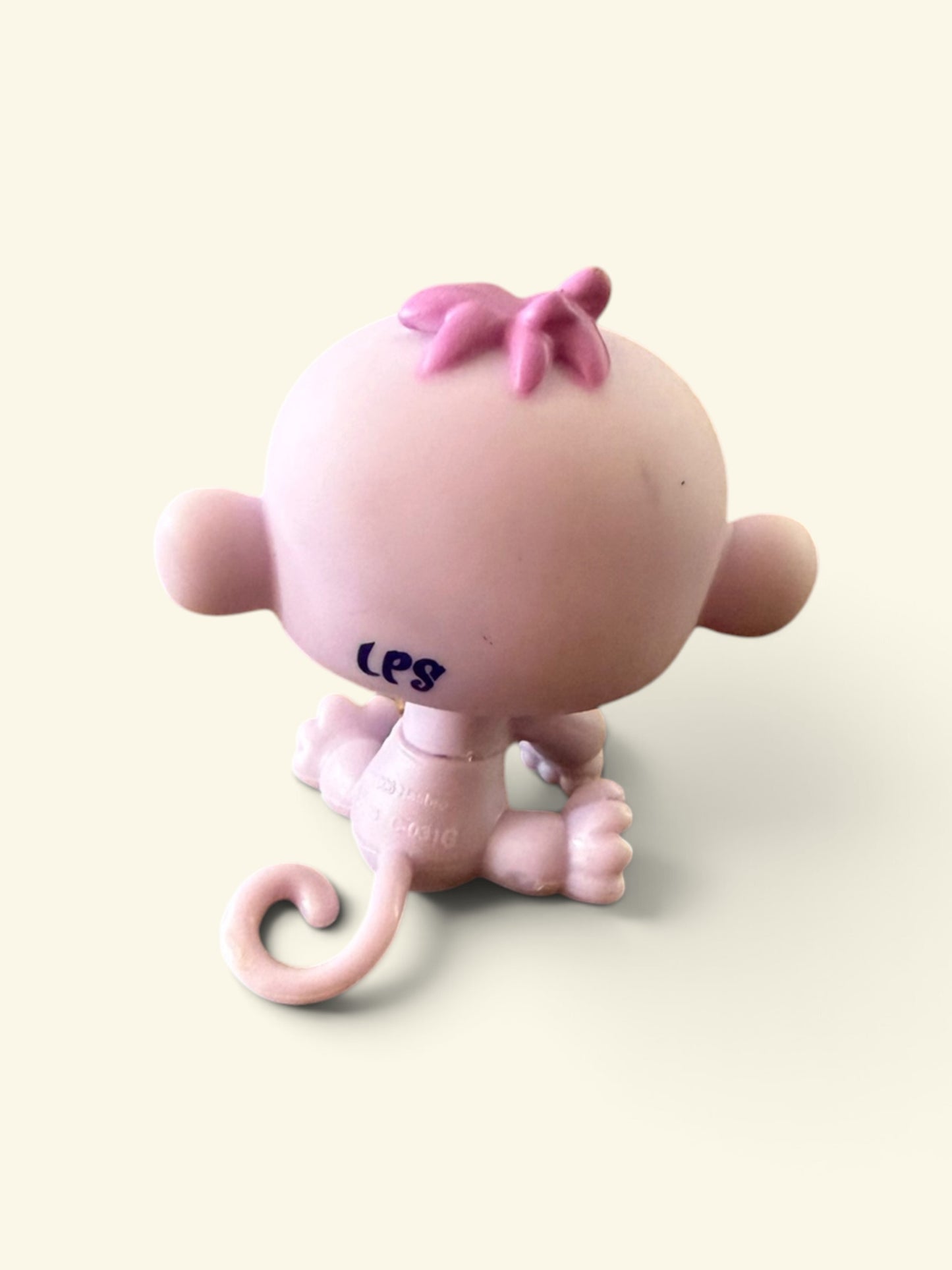 A small pink toy monkey figurine shown from the back, with a curly tail, round head, and small ears, photographed on a plain light cream background.