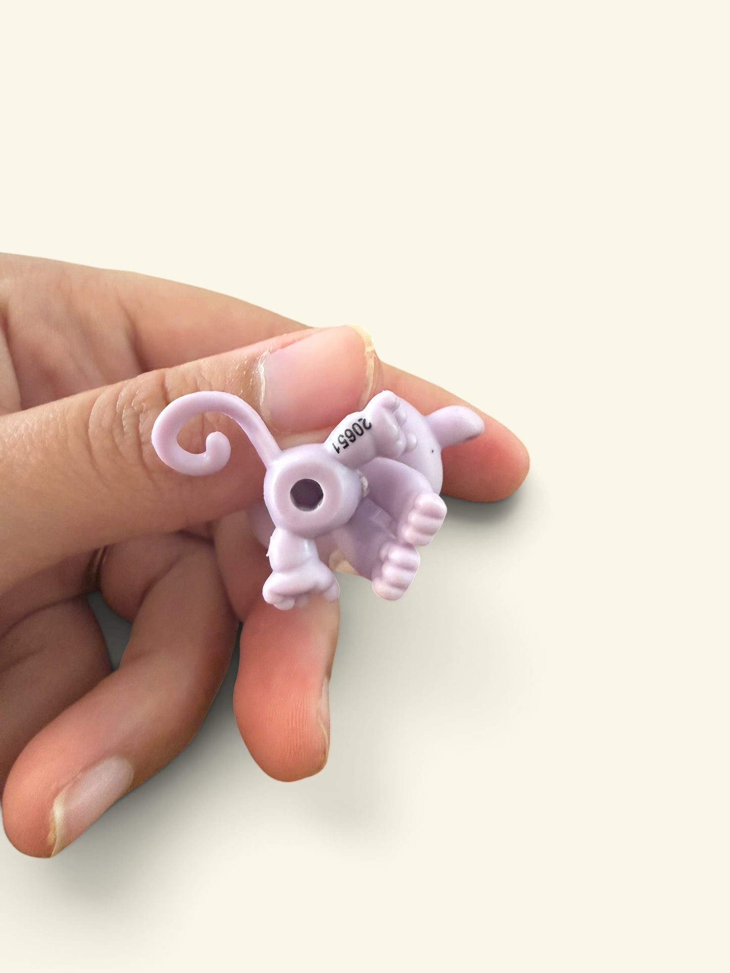 A small lavender plastic toy or accessory with a curled tail and small printed number, held between a person's fingers against a plain light beige background.