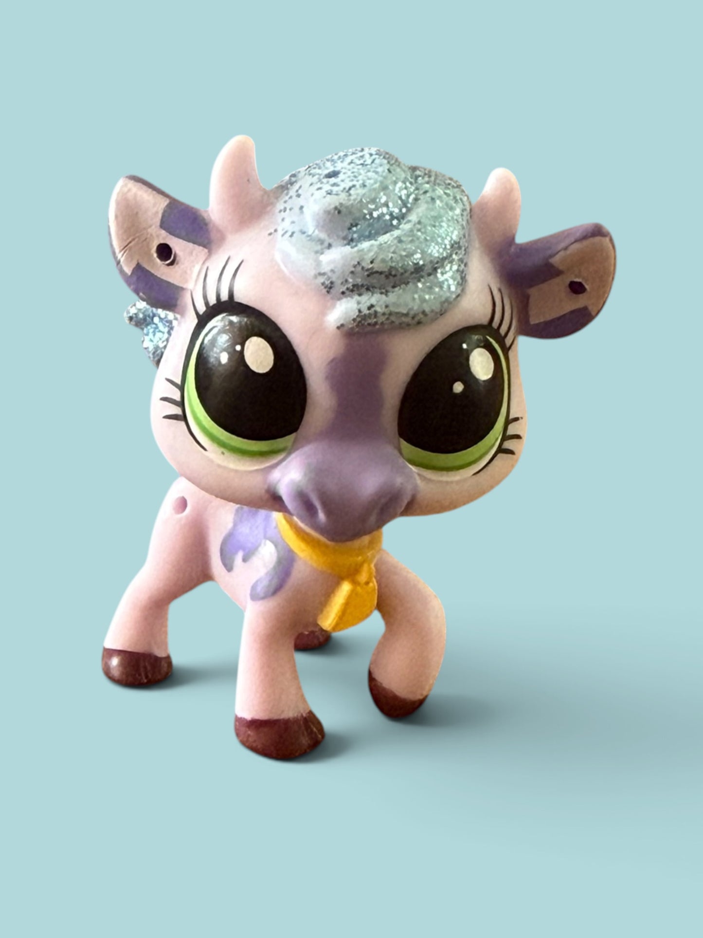 A small stylized toy animal figure resembling a cow or cow with oversized glossy eyes, a lavender face and ears, pink body, purple markings, a glittery silver-gray mane, and a yellow neck ribbon, photographed against a plain light blue background.