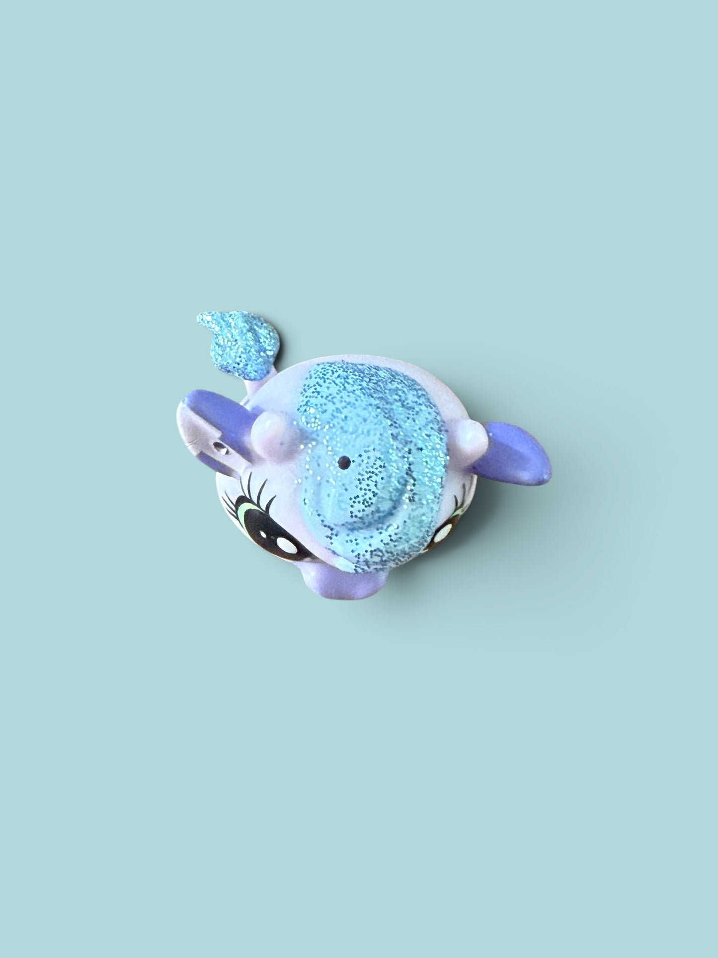 A small plush toy shaped like a round, cartoonish creature with a glittery light blue body, lavender-purple ears and tail, and large black-and-white eyes, centered on a pale blue background.
