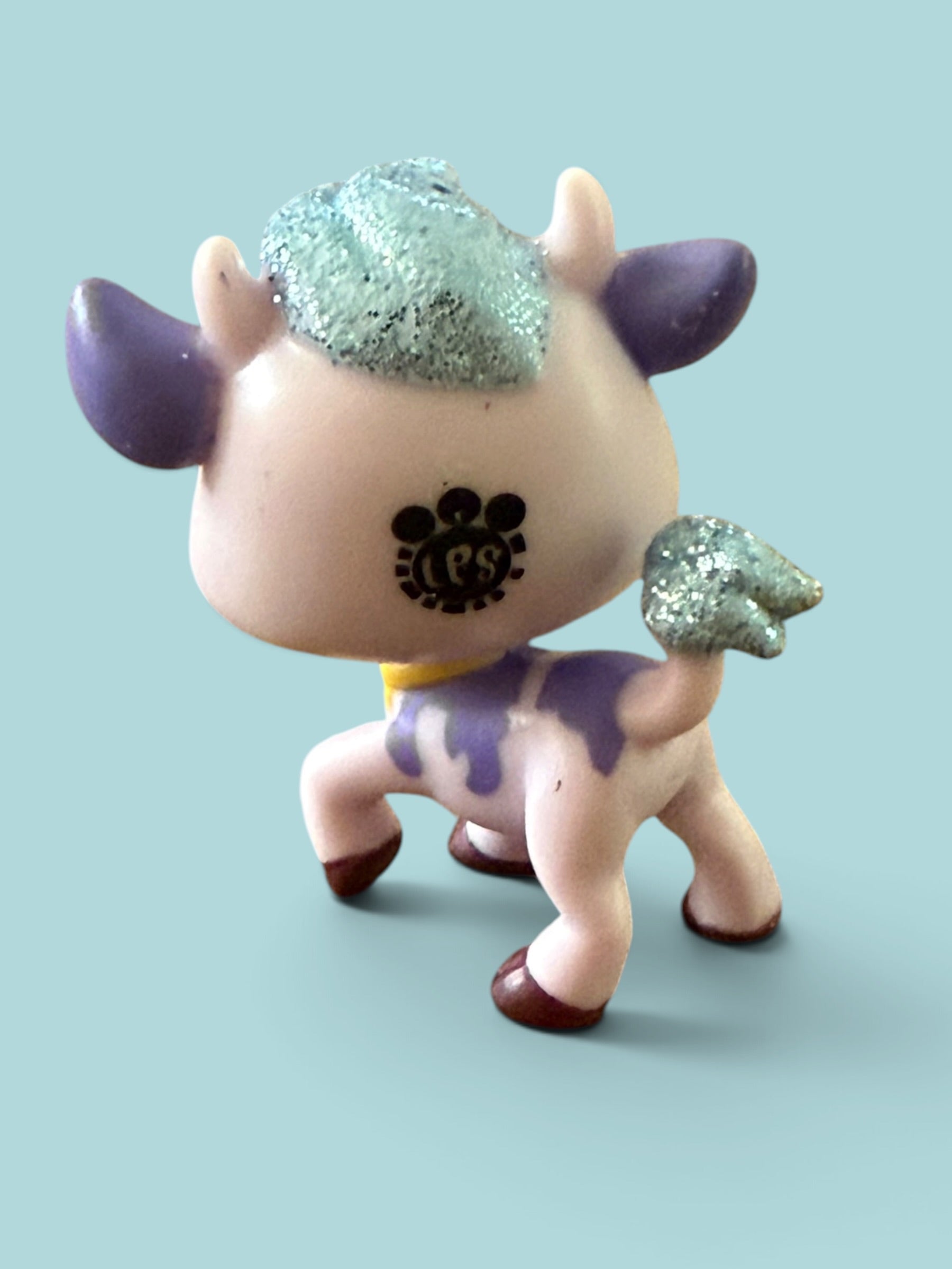 A small stylized cow figurine with a pale pink body, purple horns and body markings, glittery mint green hair, and dark brown hooves, shown against a solid light blue background.