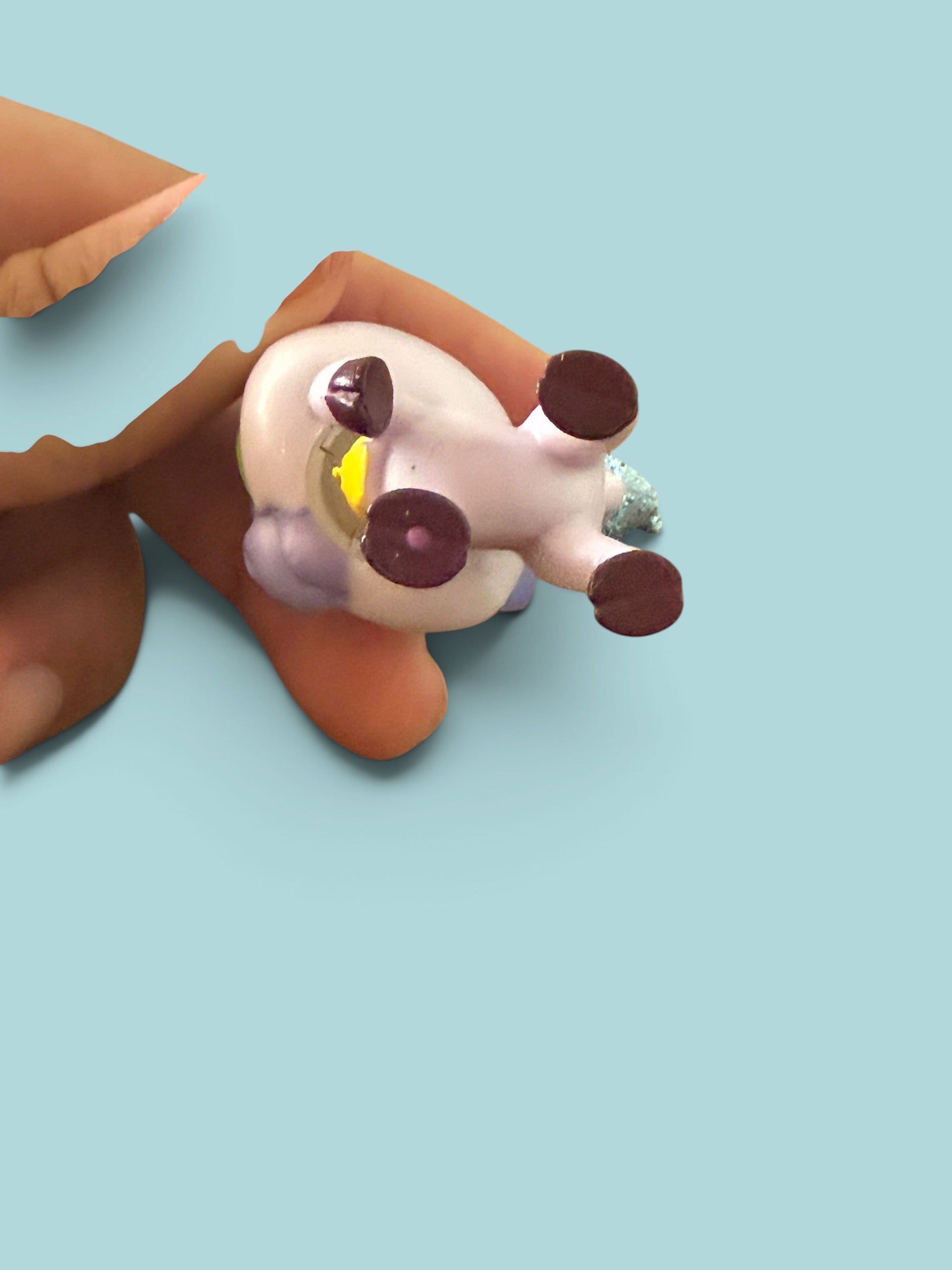 A small white plush toy with dark brown paw pads and a yellow accent on its side, held in a person's hand against a solid light blue background.