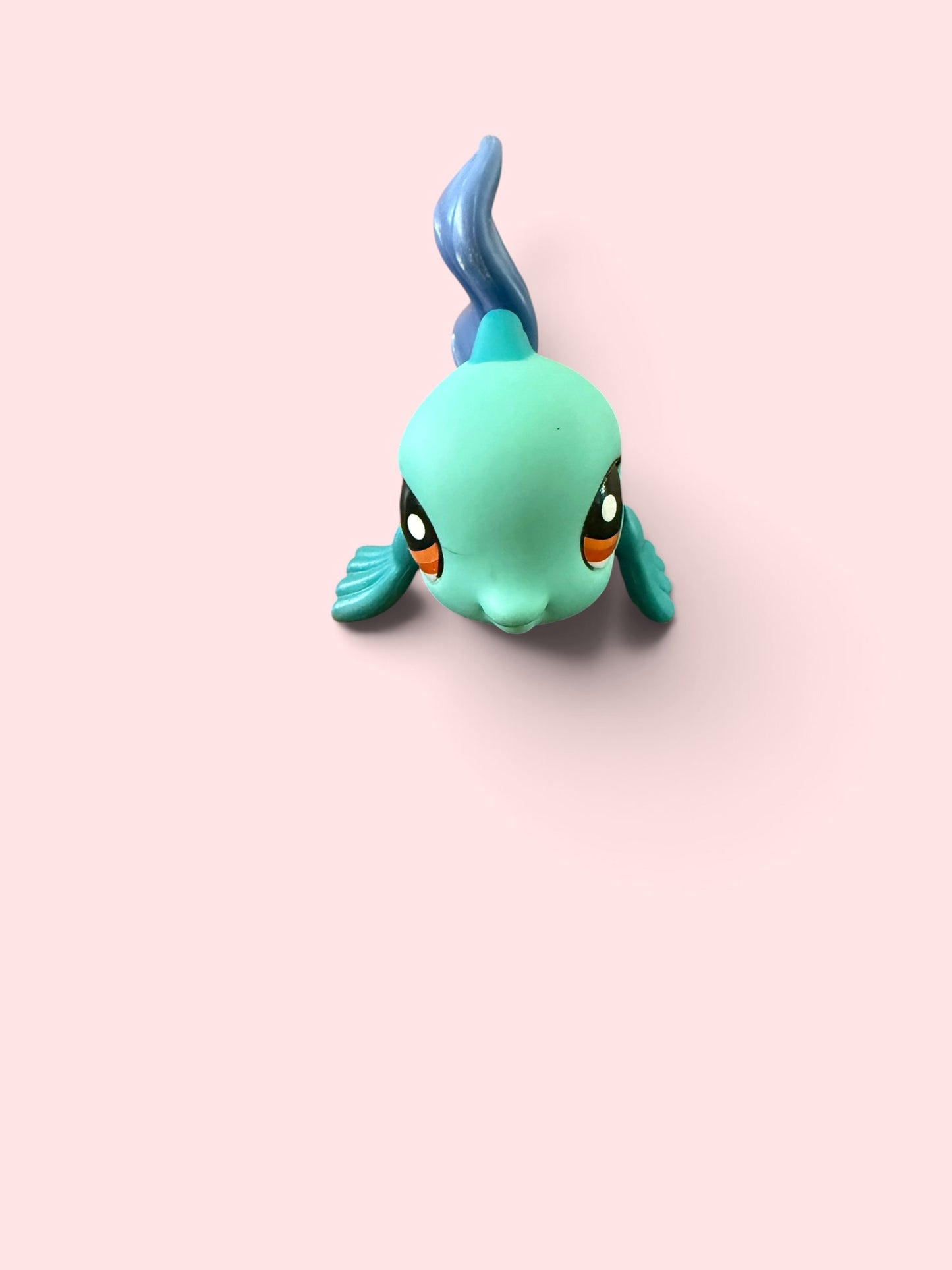 A small stylized fish figurine in mint green and blue, shown from above on a solid pale pink background.