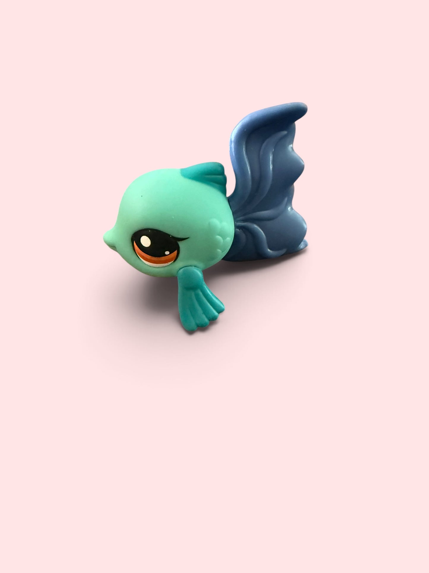 A small stylized fish figurine with a mint green head, dark blue tail and fin, and a large orange-and-black eye, shown on a plain pale pink background.
