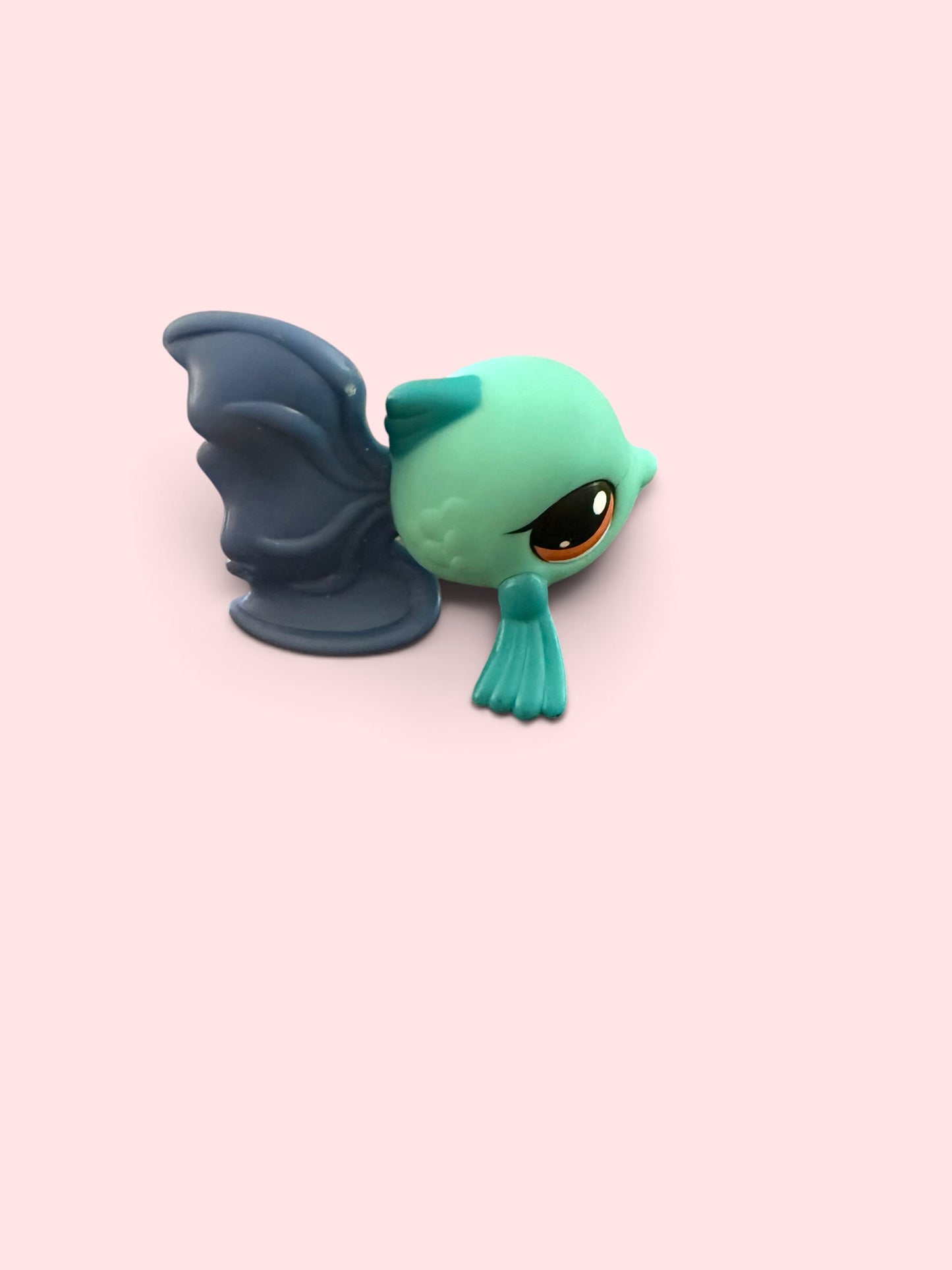 A small stylized toy figure resembling a cute fish or sea creature with a mint green head, dark blue wings, and a matching dark blue tail, shown on a plain pale pink background.