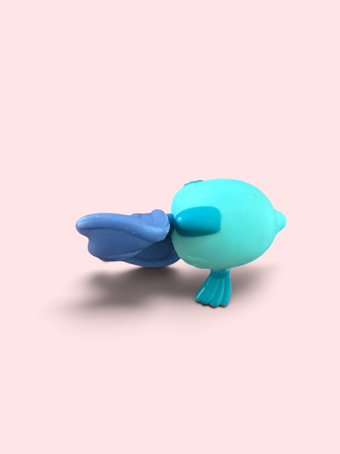 A small stylized fish figurine in bright turquoise and blue, shown from the side on a soft pink background.