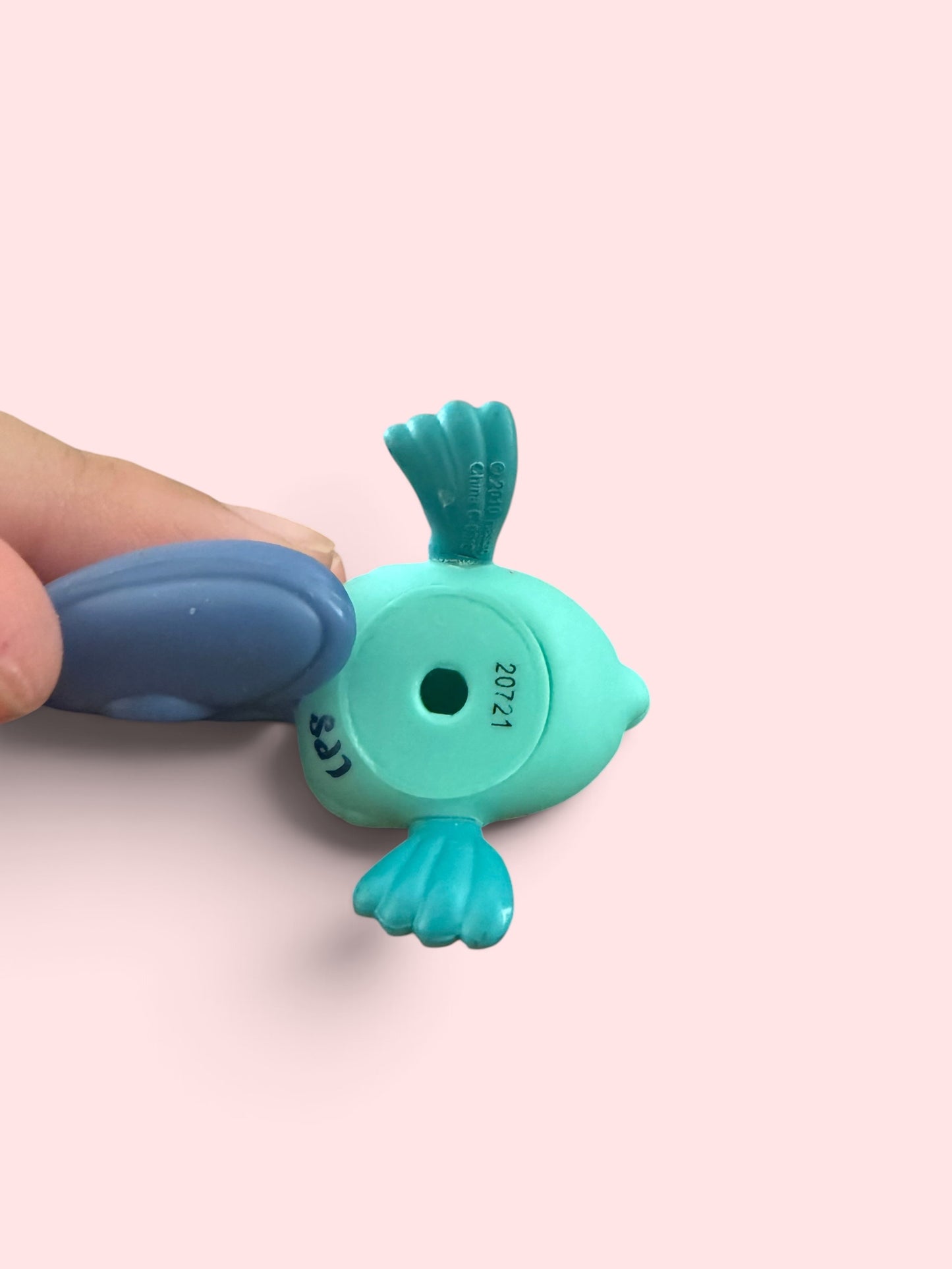 A close-up of a small turquoise fish-shaped object being held by a human hand against a soft pink background. The object has a circular hole in the center and printed black text reading '20721' and 'LAL'.