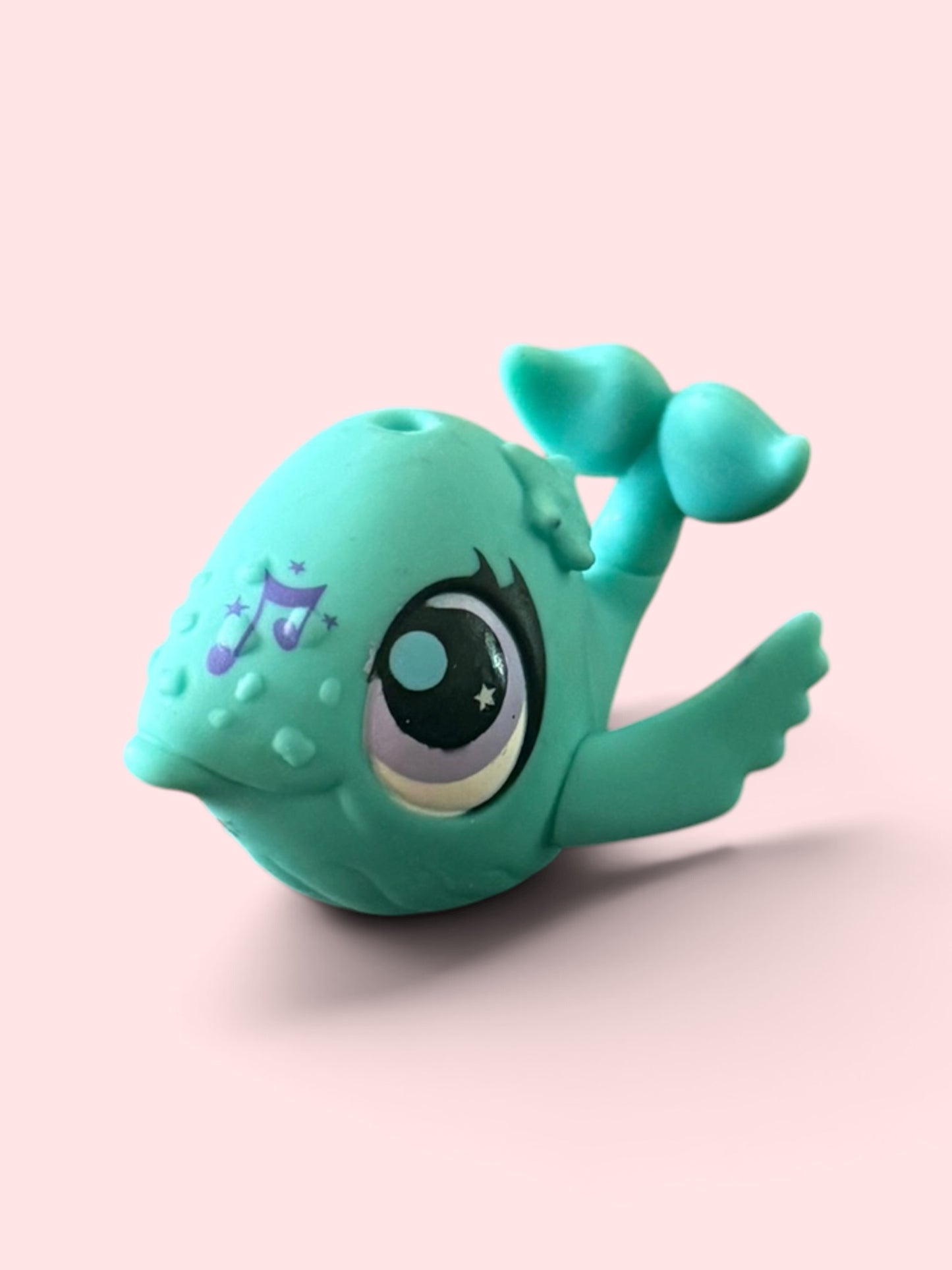 A small teal whale-shaped novelty item with large cartoon eyes, a purple musical note and star design on its forehead, and a tail fin, shown on a soft pink background.