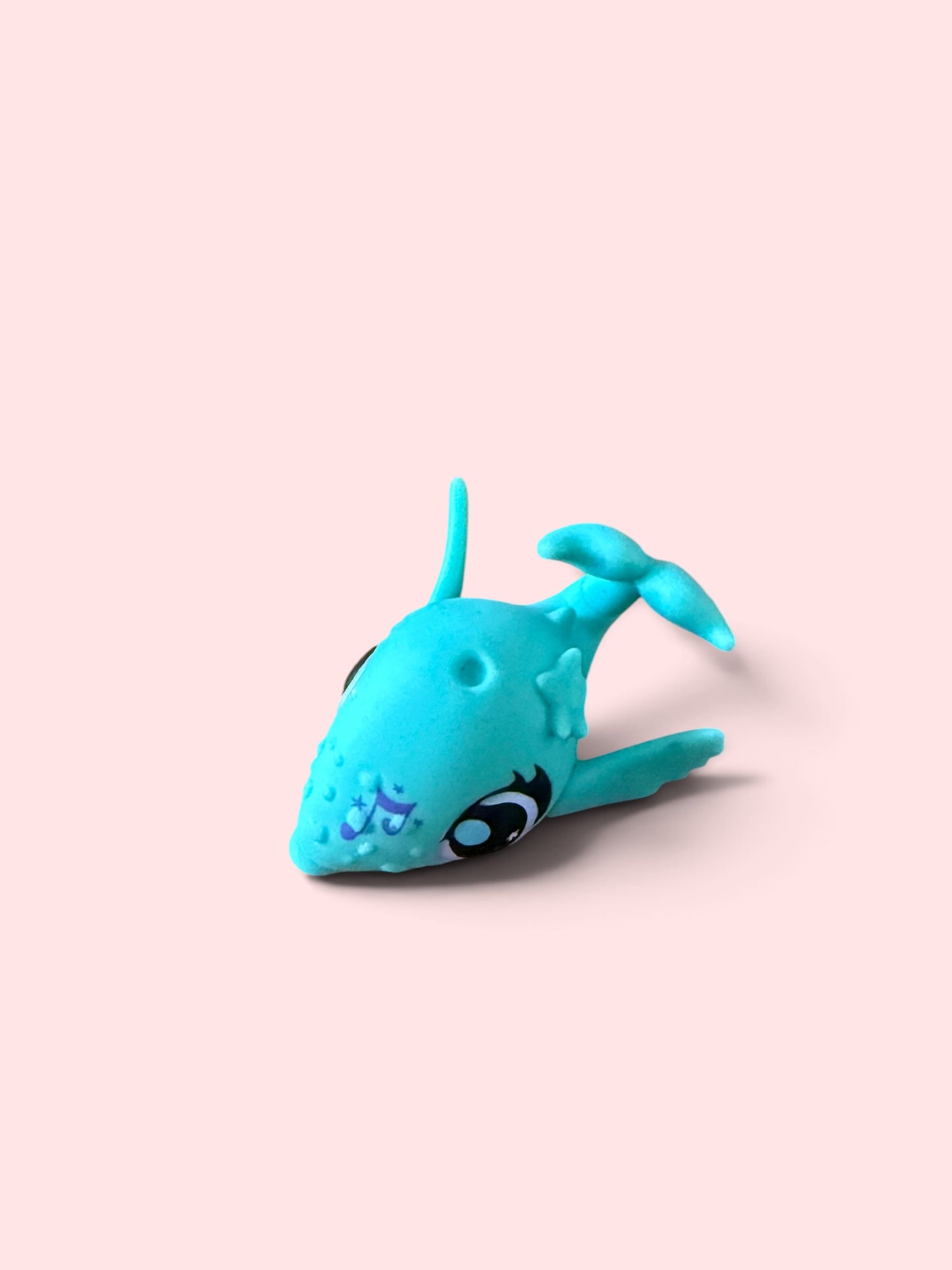 A small turquoise whale-shaped toy or accessory with large cartoon eyes and textured details, photographed on a soft light pink background.