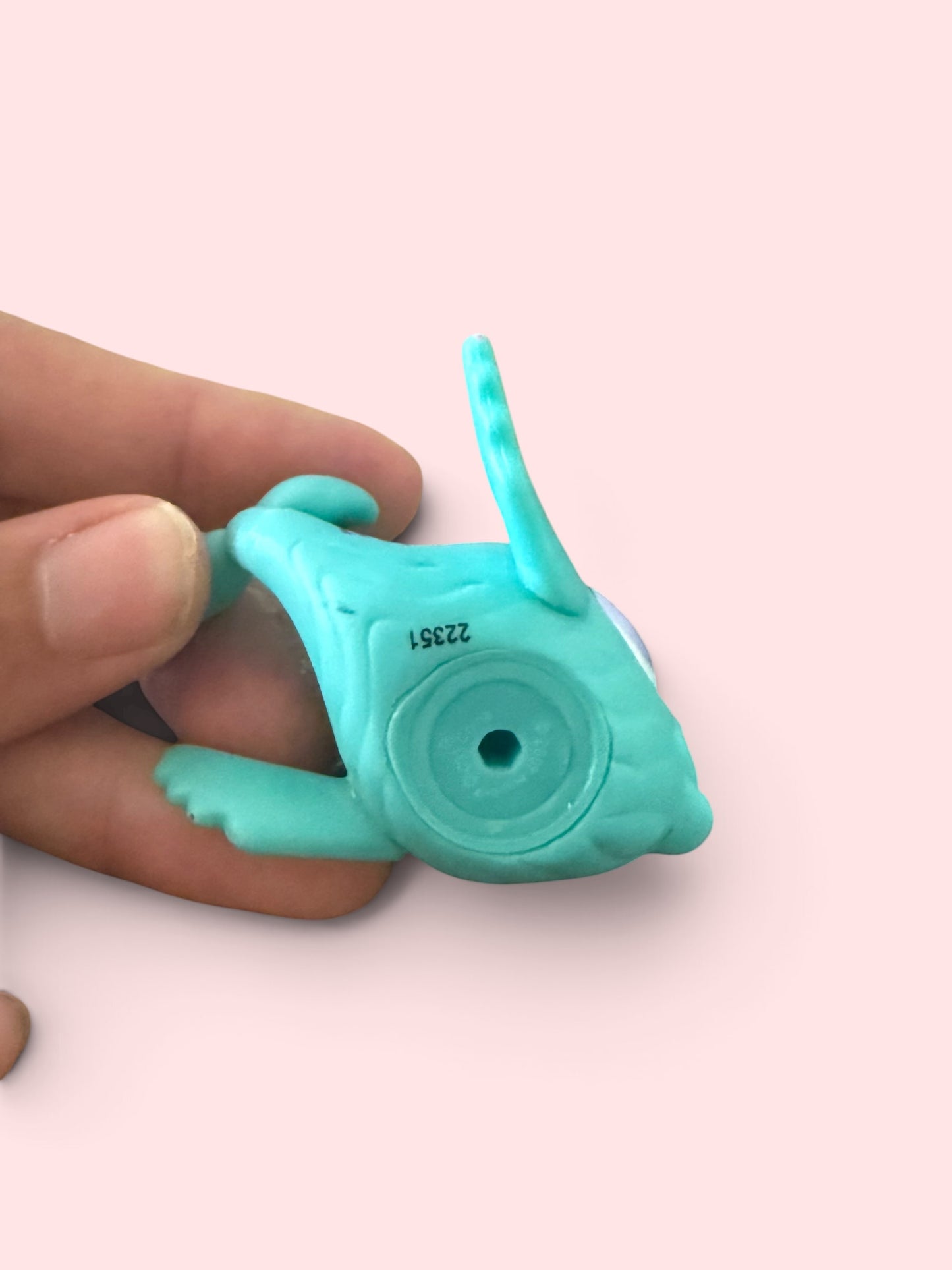 A small turquoise plastic toy or accessory being held between fingers against a pale pink background.