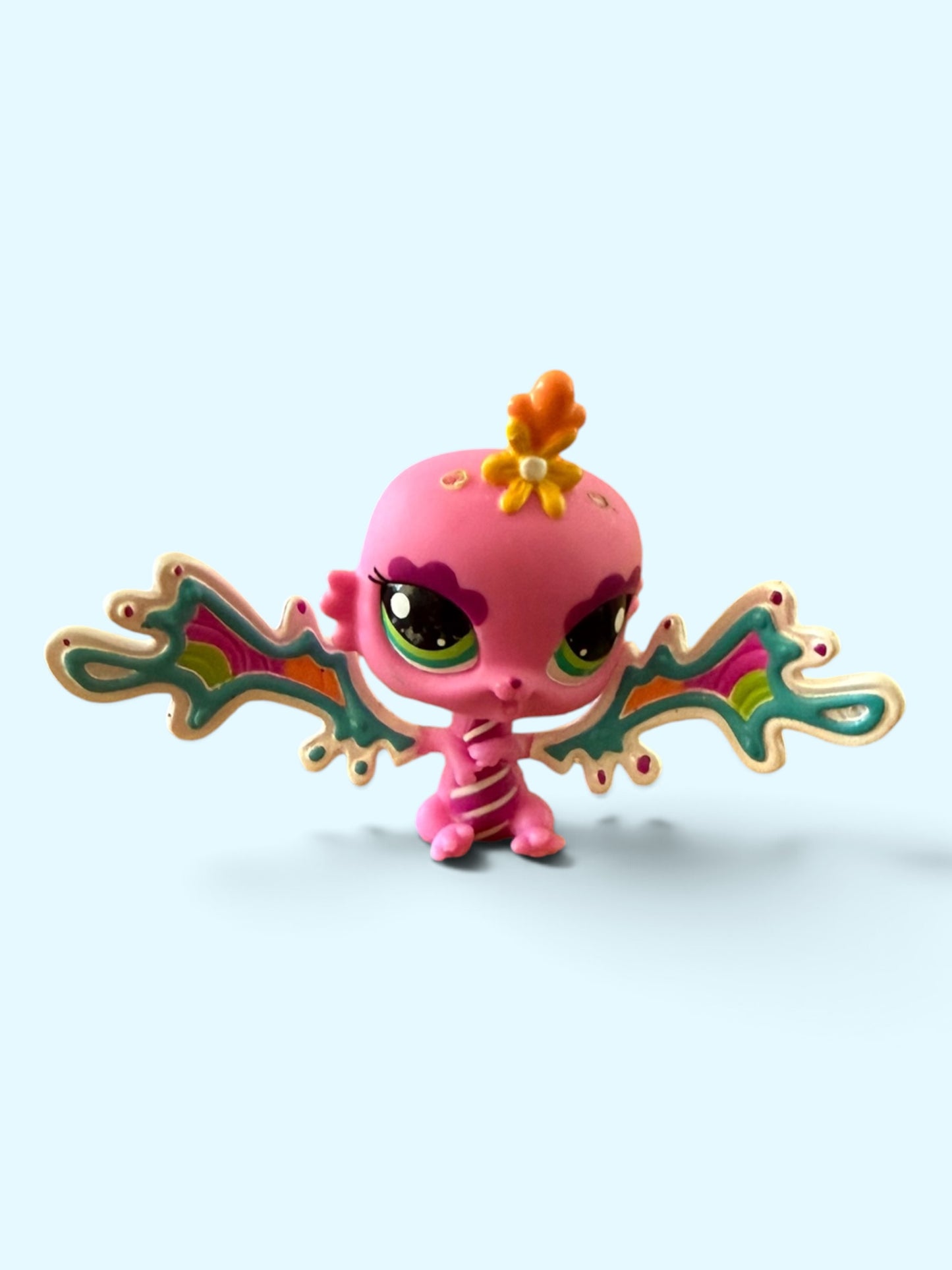 A small pink fantasy toy creature with large green eyes, a yellow flower on its head, and colorful patterned wings, centered on a pale blue background.