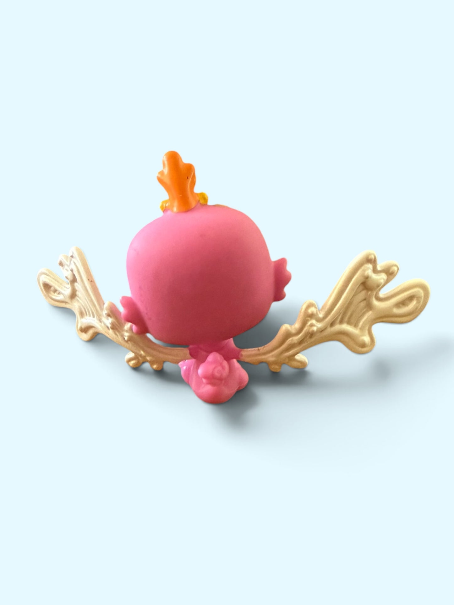 A small pink fantasy figurine with an orange flame on its head and ornate gold wings, shown from the back on a light blue background.