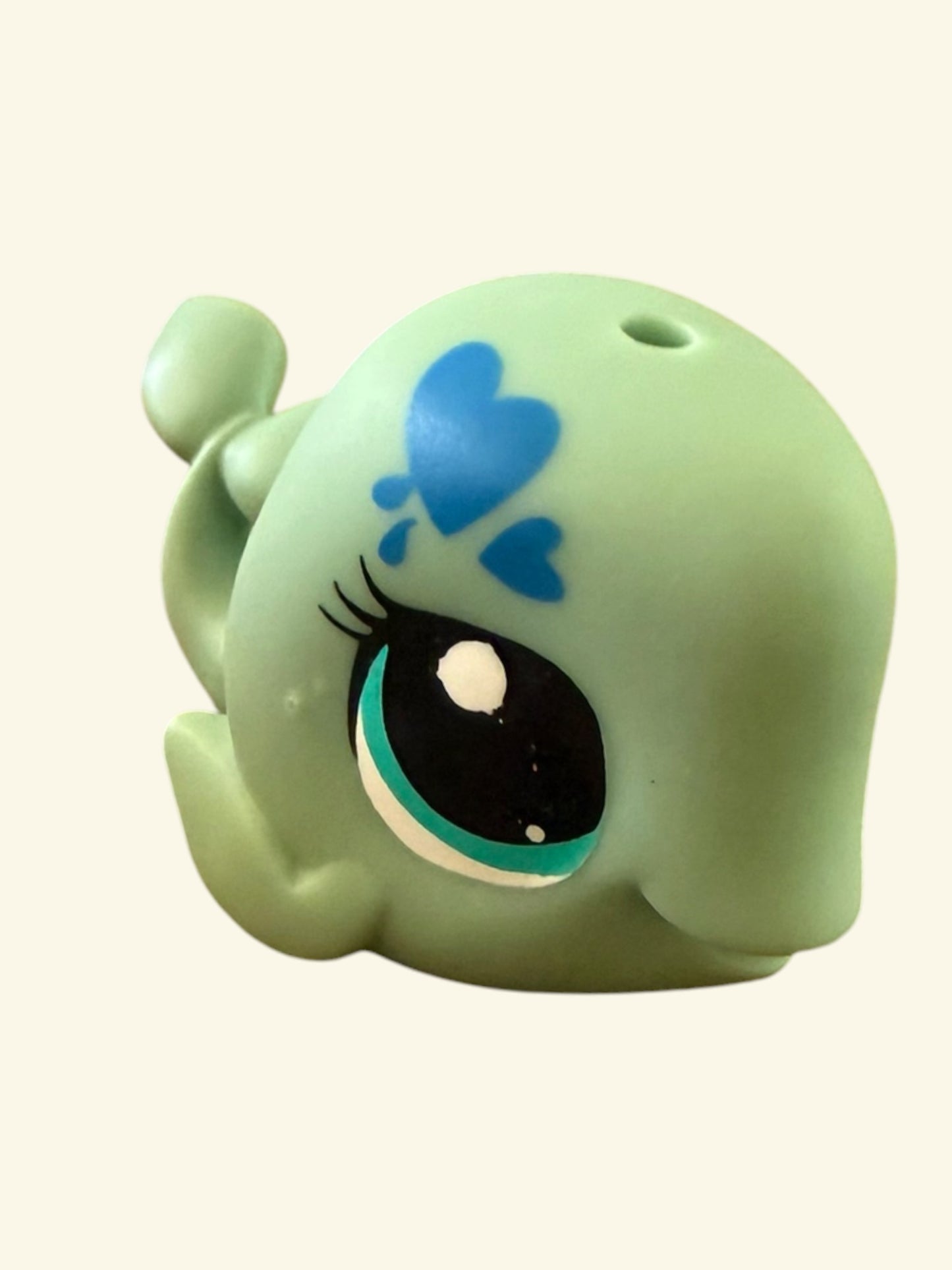 A small mint green whale-shaped plastic toy or figurine with a large black eye, teal iris, and blue heart details on the forehead, shown against a plain off-white background.