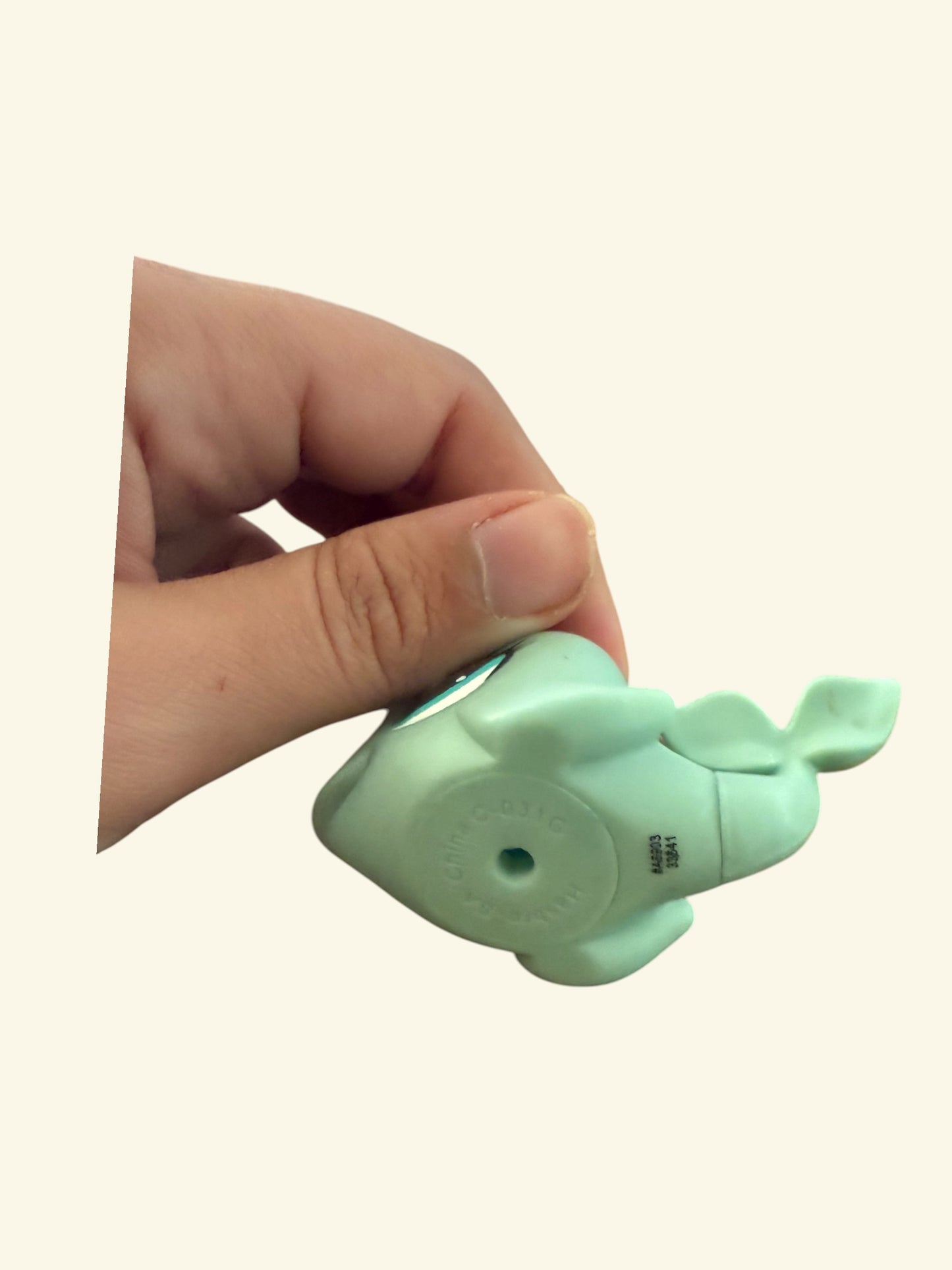 A hand holding a mint green whale-shaped bottle or container against a plain light beige background.