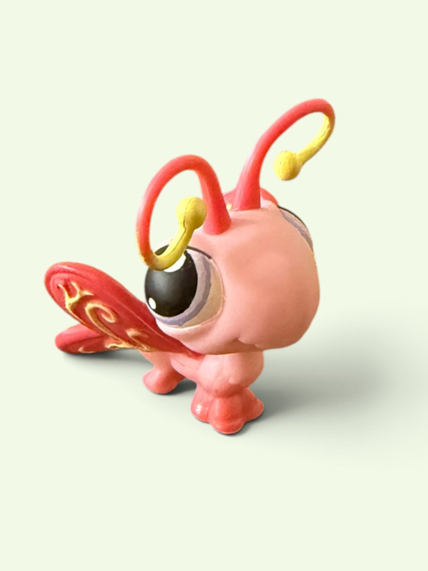 A stylized pink cartoon creature figurine with large black eyes, curved red antennae tipped in yellow, and a red wing-like element with gold swirls, shown on a pale green background.