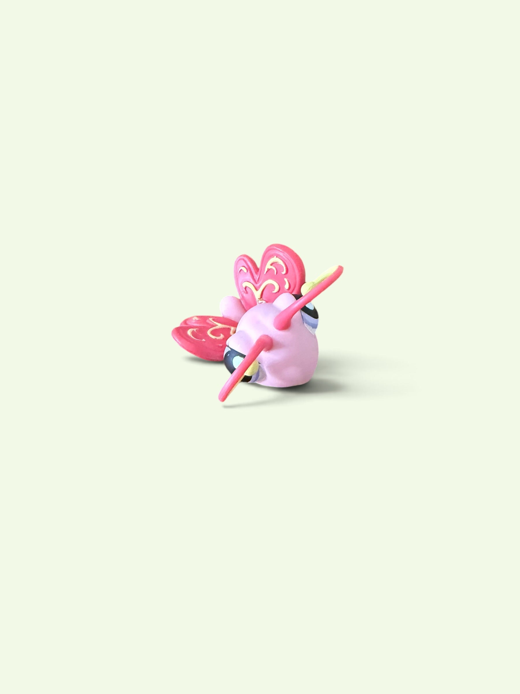 A small pink decorative figurine or ornament shaped like a stylized character with pink wings and a pink staff, shown on a plain pale cream background.