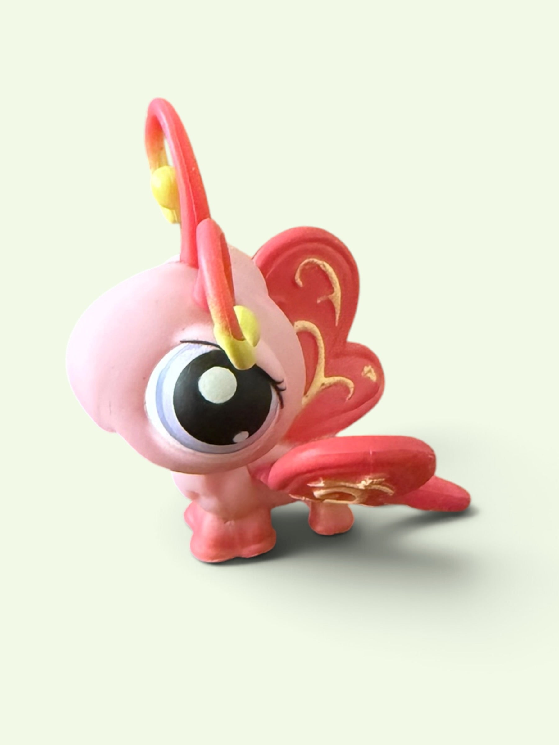 A small pink fantasy creature toy with a large glossy eye, red wings with gold detailing, and yellow accents, shown on a plain light background.