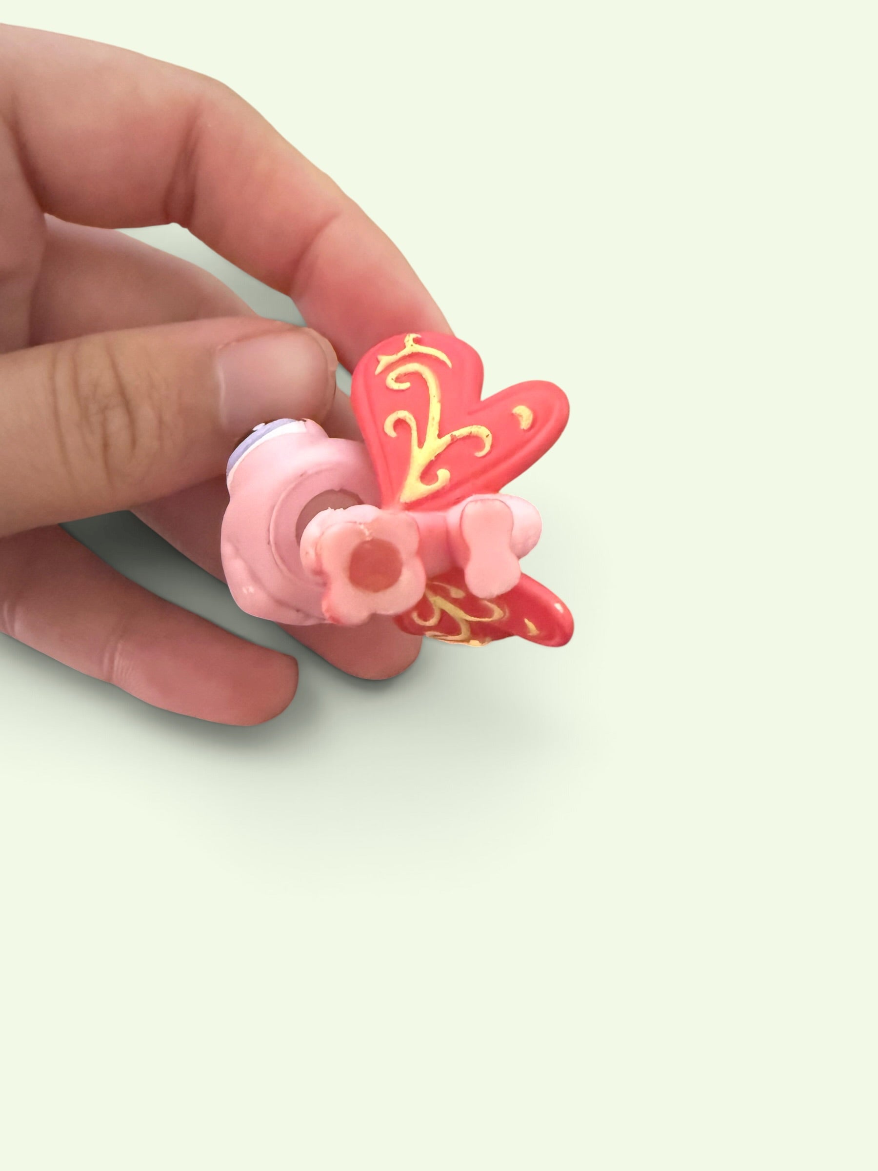 A hand holding a small pink and red butterfly  figurine with gold decorative details against a pale green background.