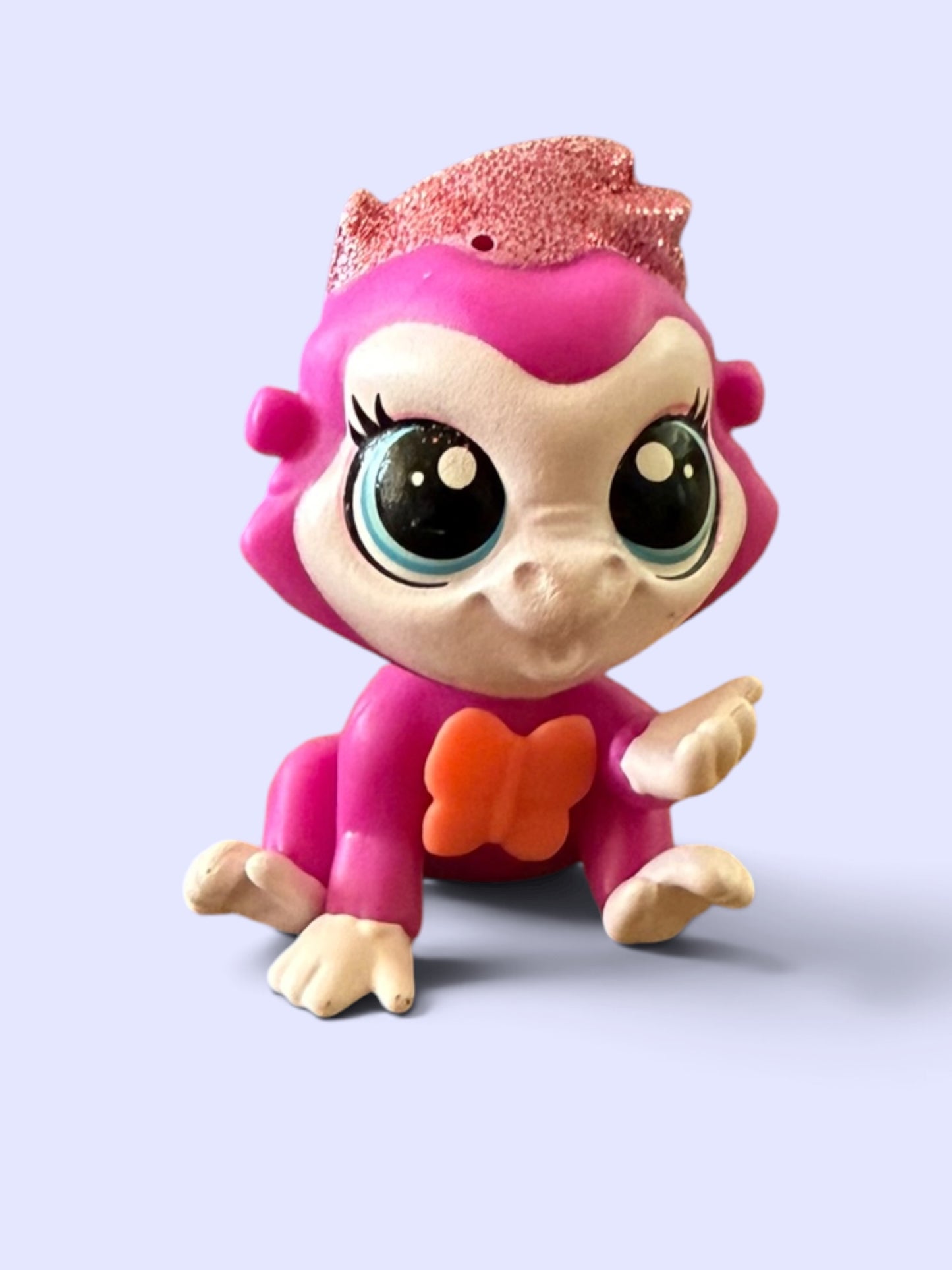 A small pink monkey toy figurine with large cartoon eyes, cream-colored face and hands, a glittery pink crown, and an orange bow on the chest, shown against a pale lavender background.