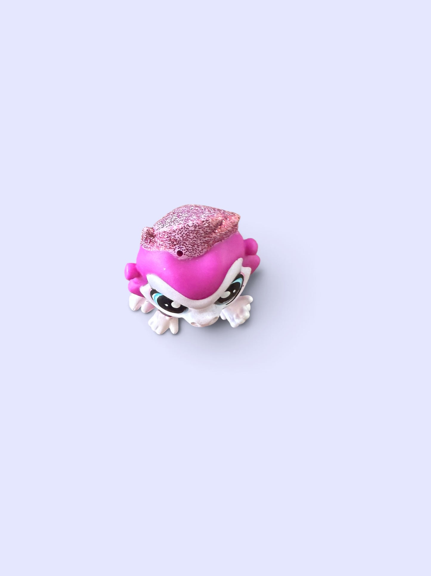 A small stylized toy figure with a pink glittery hat, bright pink hair, and a white body, centered on a pale lavender background.